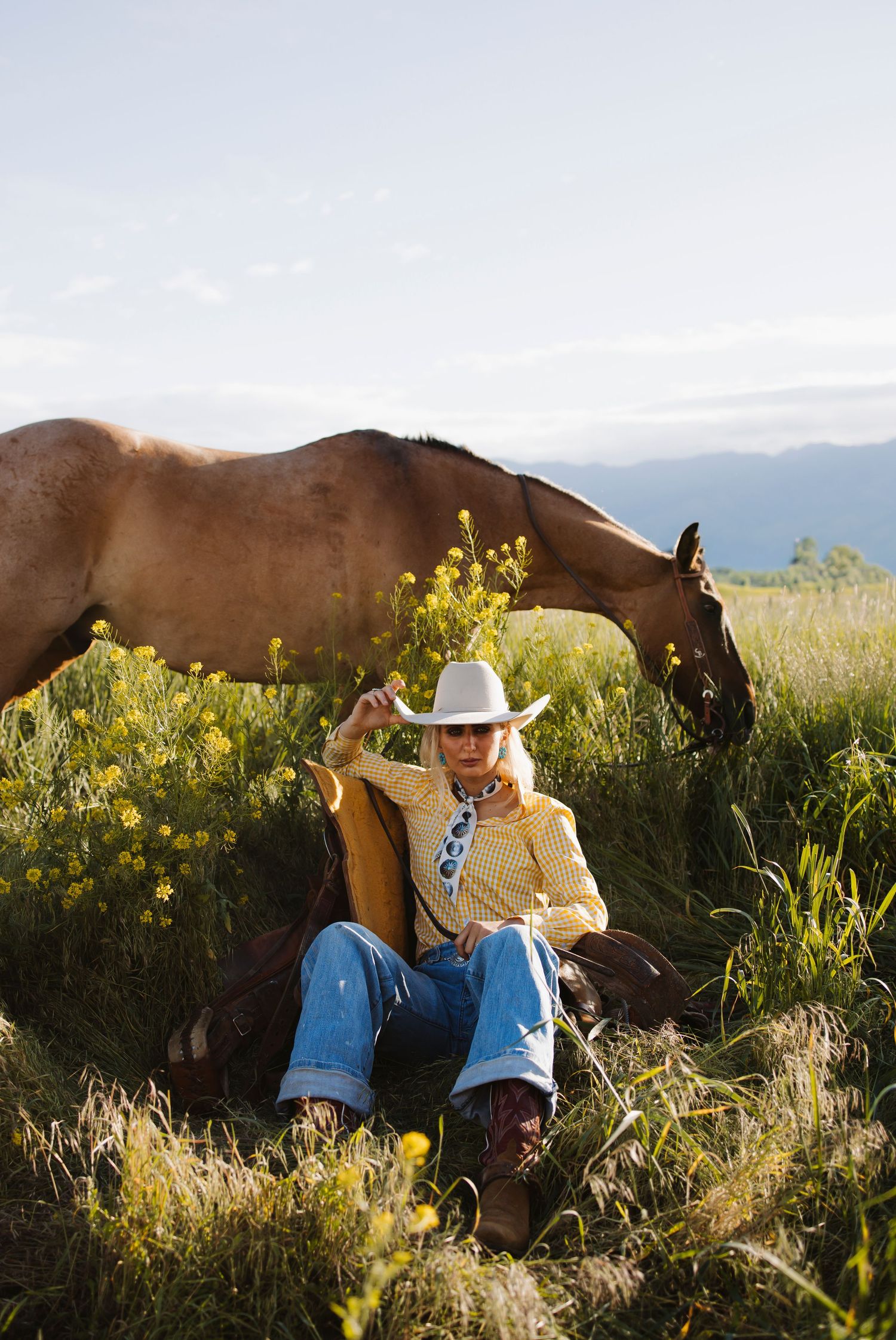 A blonde woman in a cowboy hat, yellow shirt, and jeans sits in a wildflower field beside a grazing brown horse.