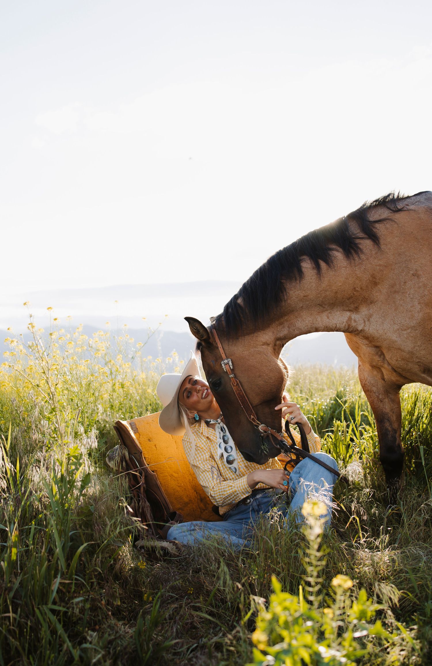 A cowboy sits in a sunny meadow bonding with a brown horse nuzzling close, surrounded by wildflowers and rolling hills.