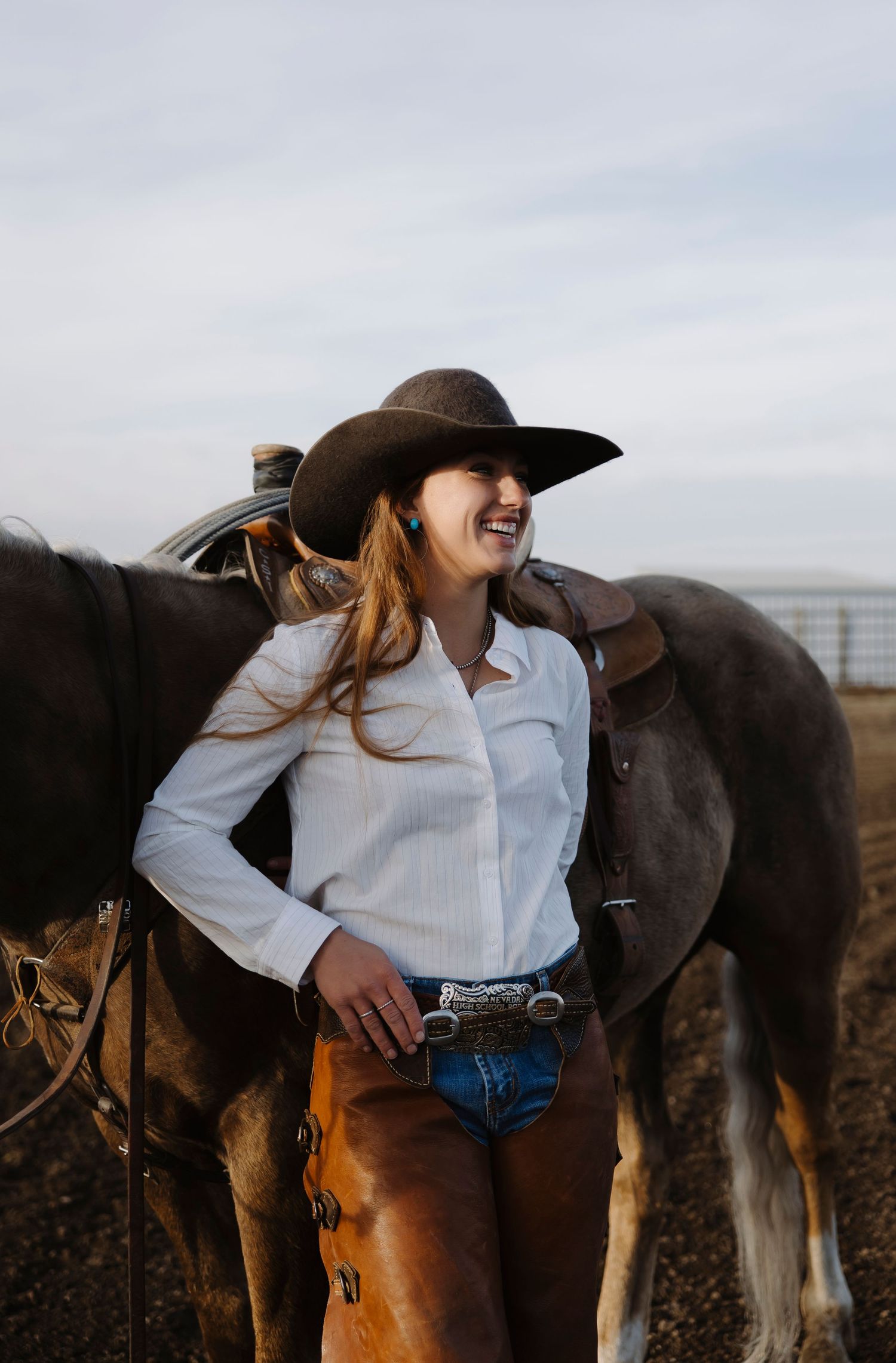 Smiling cowgirl in white shirt, brown chaps, and cowboy hat standing beside a dark horse outdoors.