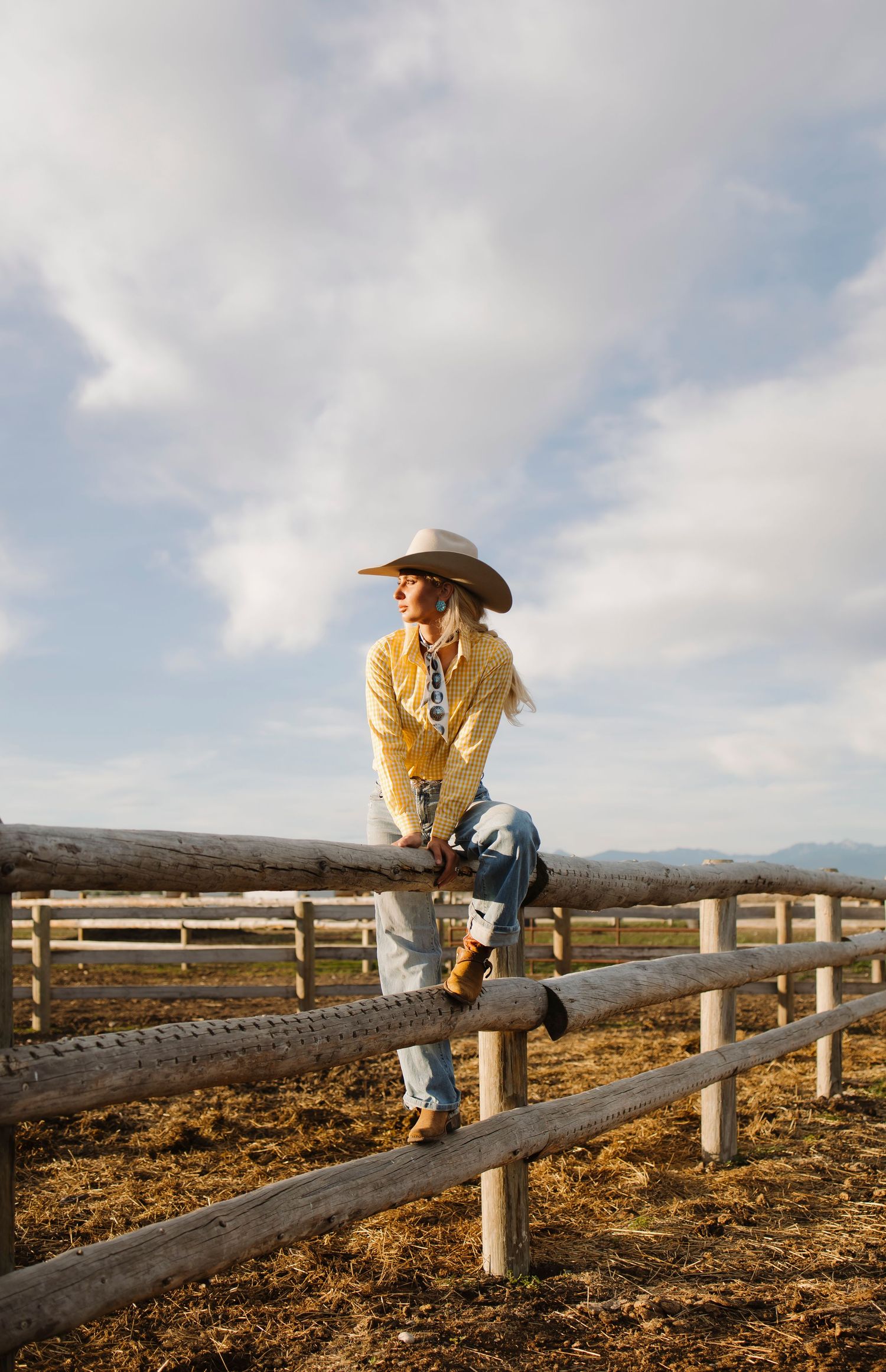 A woman in a yellow fringe jacket, cowboy hat, and jeans sits on a rustic wooden ranch fence under a cloudy sky.