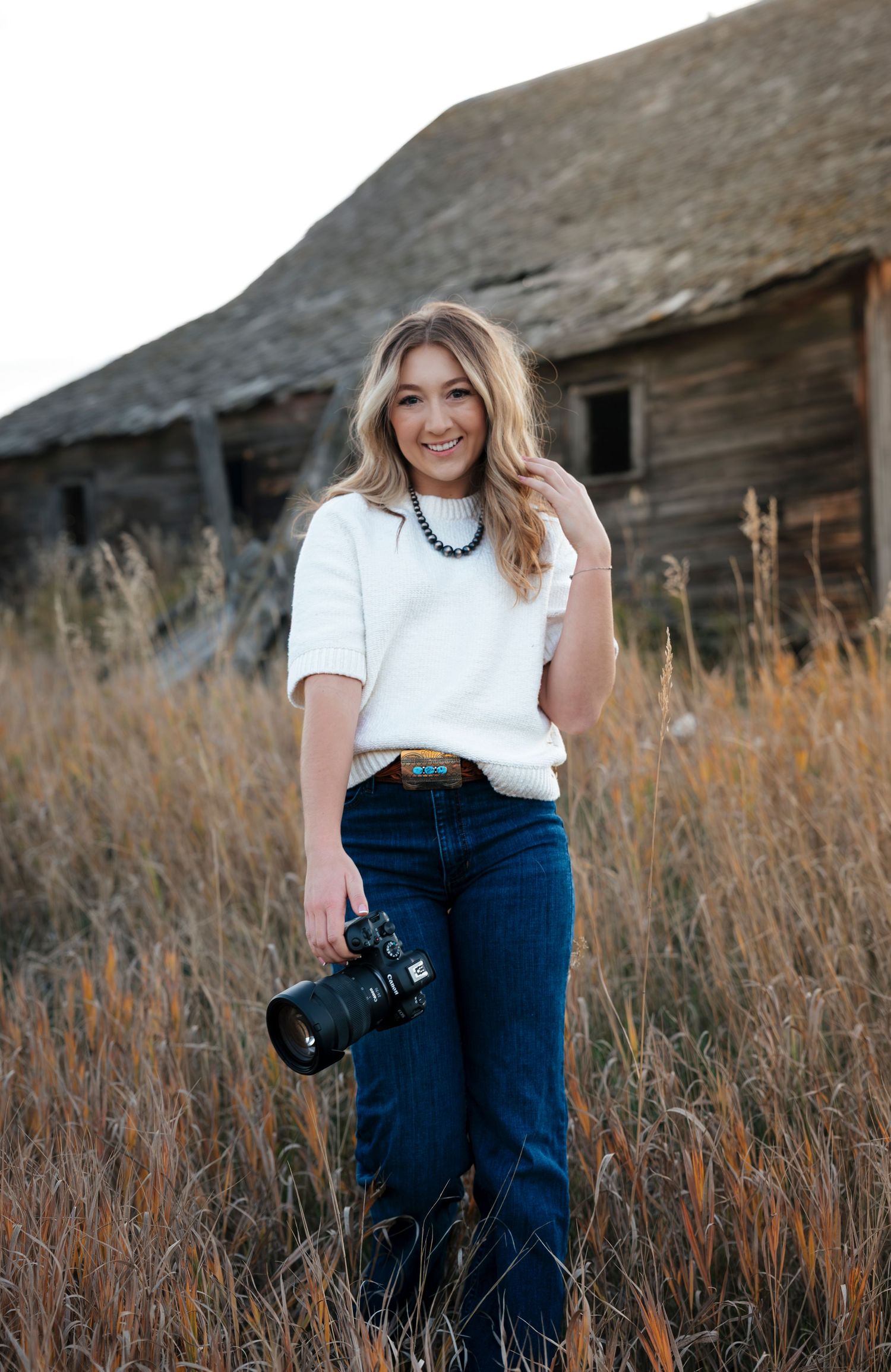 Photographer in white shirt and jeans holds camera in rustic outdoor setting with tall grass and wooden barn.