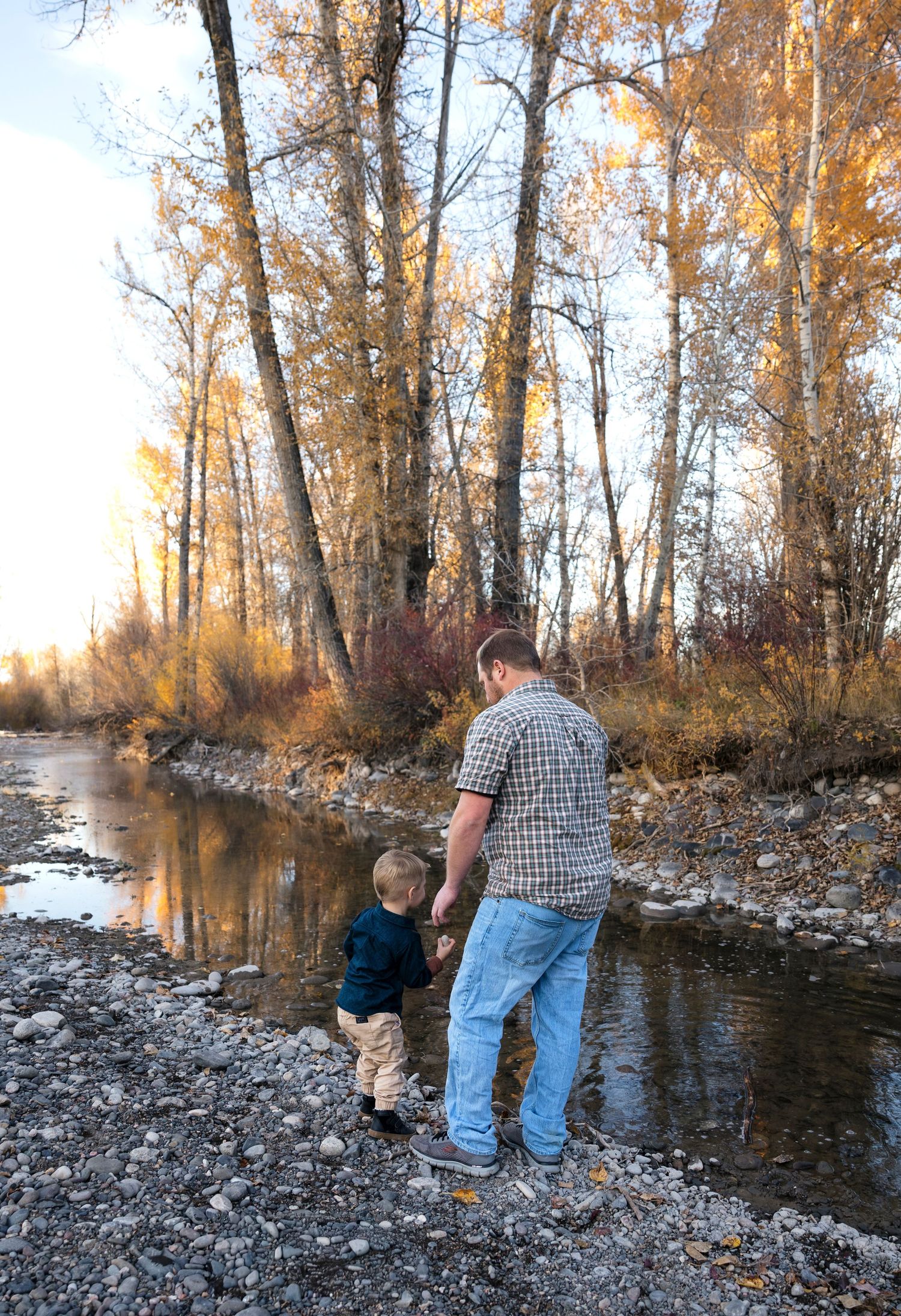 People skipping rocks into a river surrounded by autumn trees at sunset.