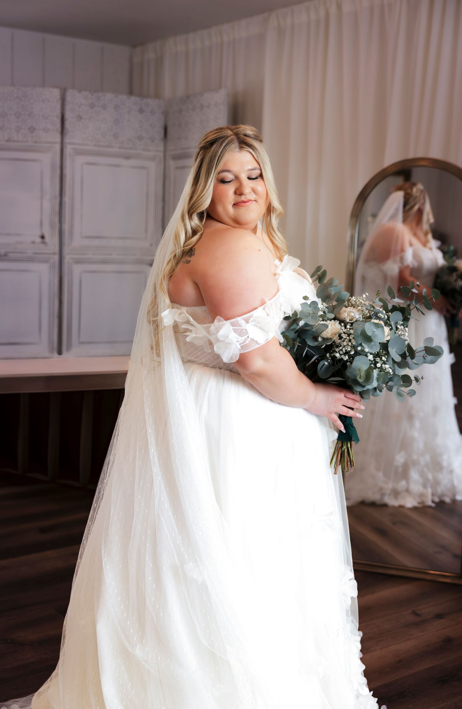 A bride in an off-shoulder white gown holds a eucalyptus bouquet in a rustic indoor setting.