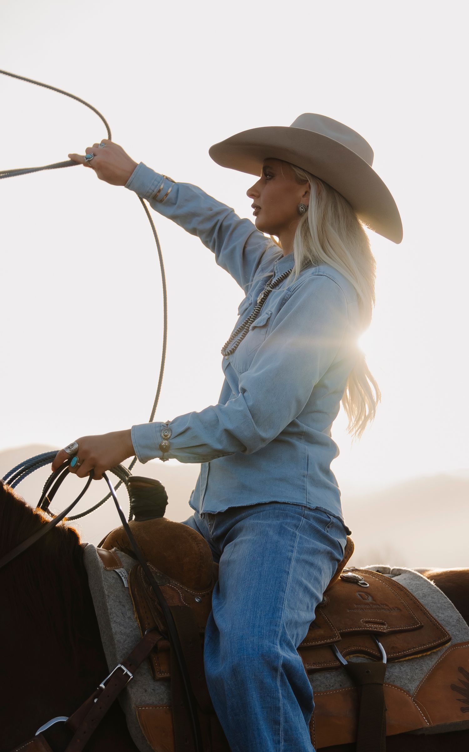 A blonde woman in a cowboy hat and denim outfit swings a lasso while riding a horse at sunset.