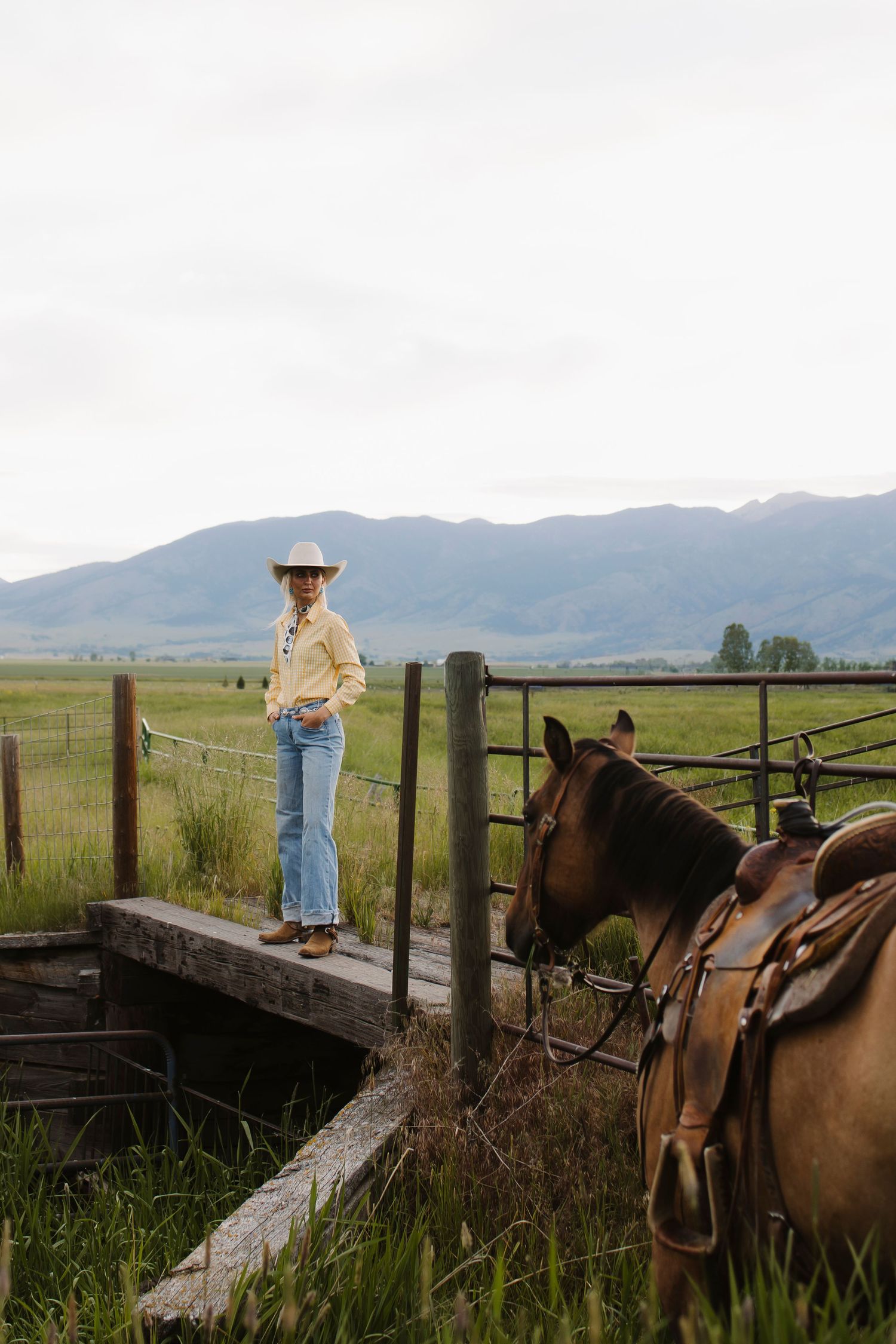 Woman in cowboy hat and jeans stands by a wooden fence with a saddled brown horse, mountains in background.