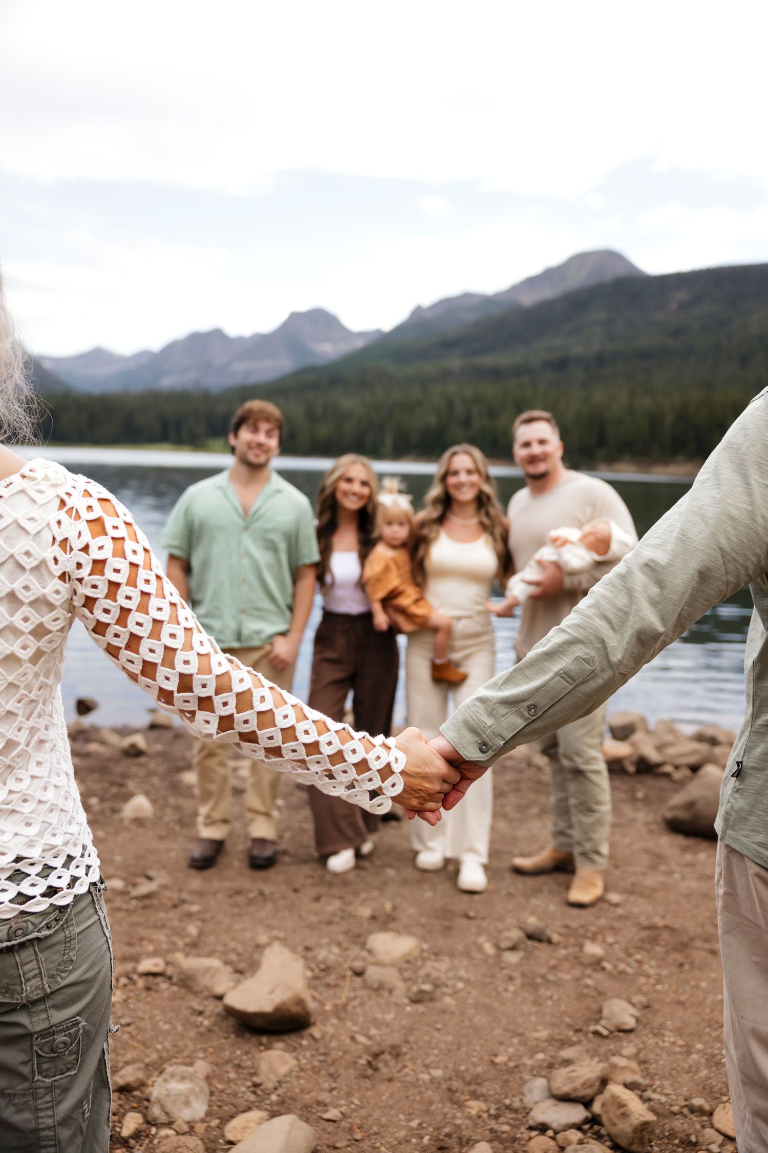Couple holding hands near a mountain lake with friends and family smiling in the background.