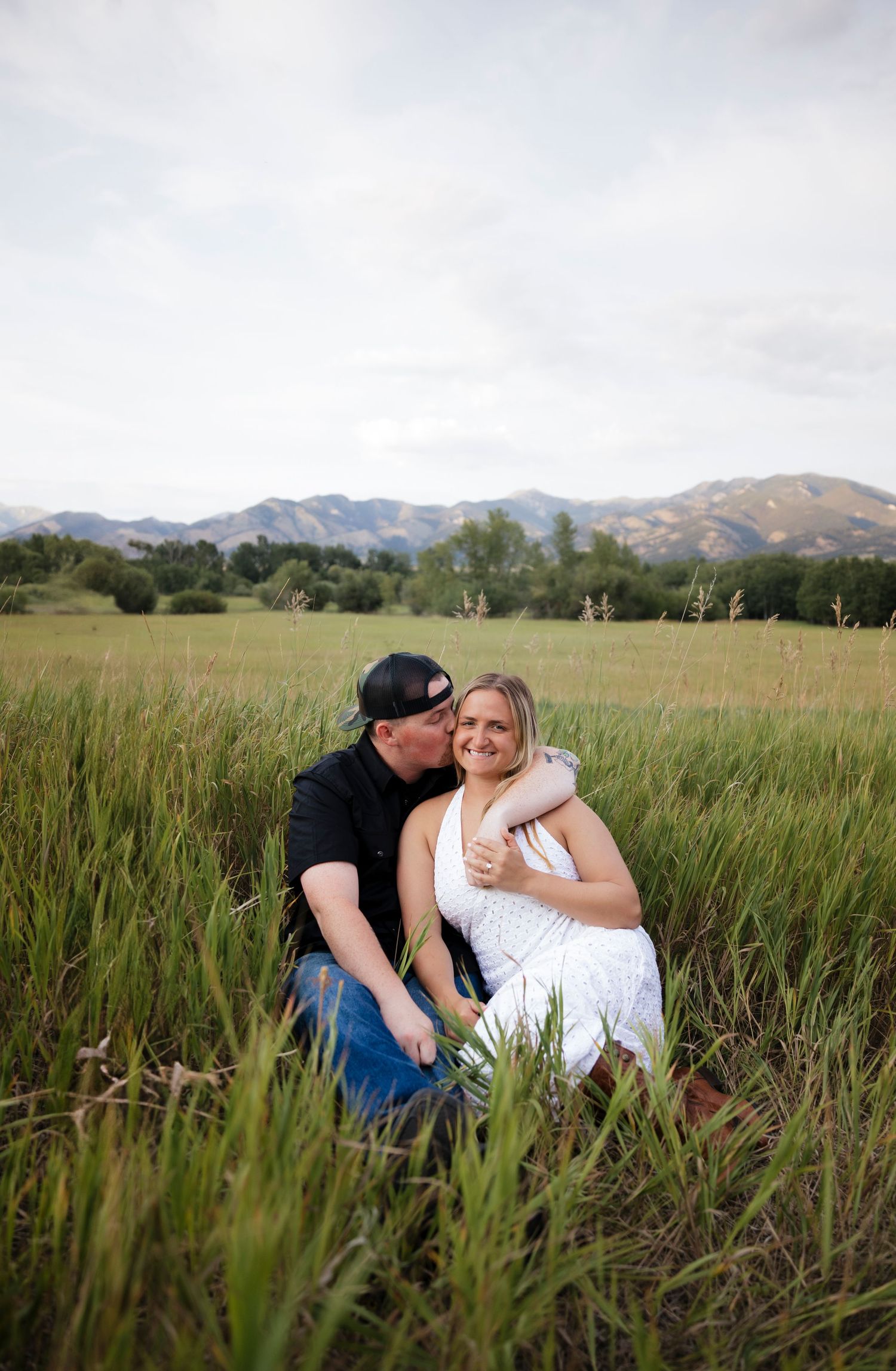 Couple sharing a tender moment while sitting in a hay field with mountains in the background.