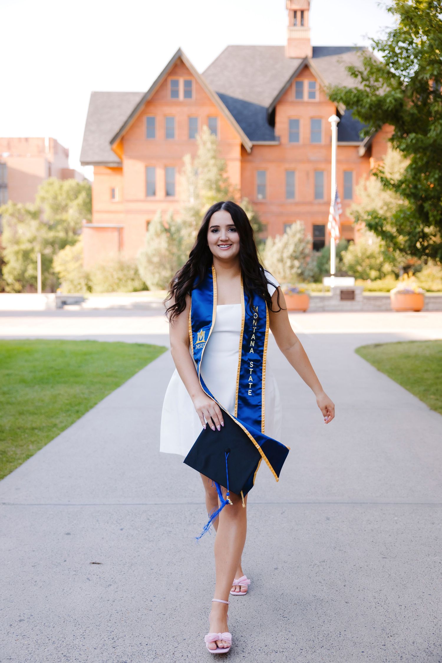 Graduate in blue stole and white dress poses on campus walkway.