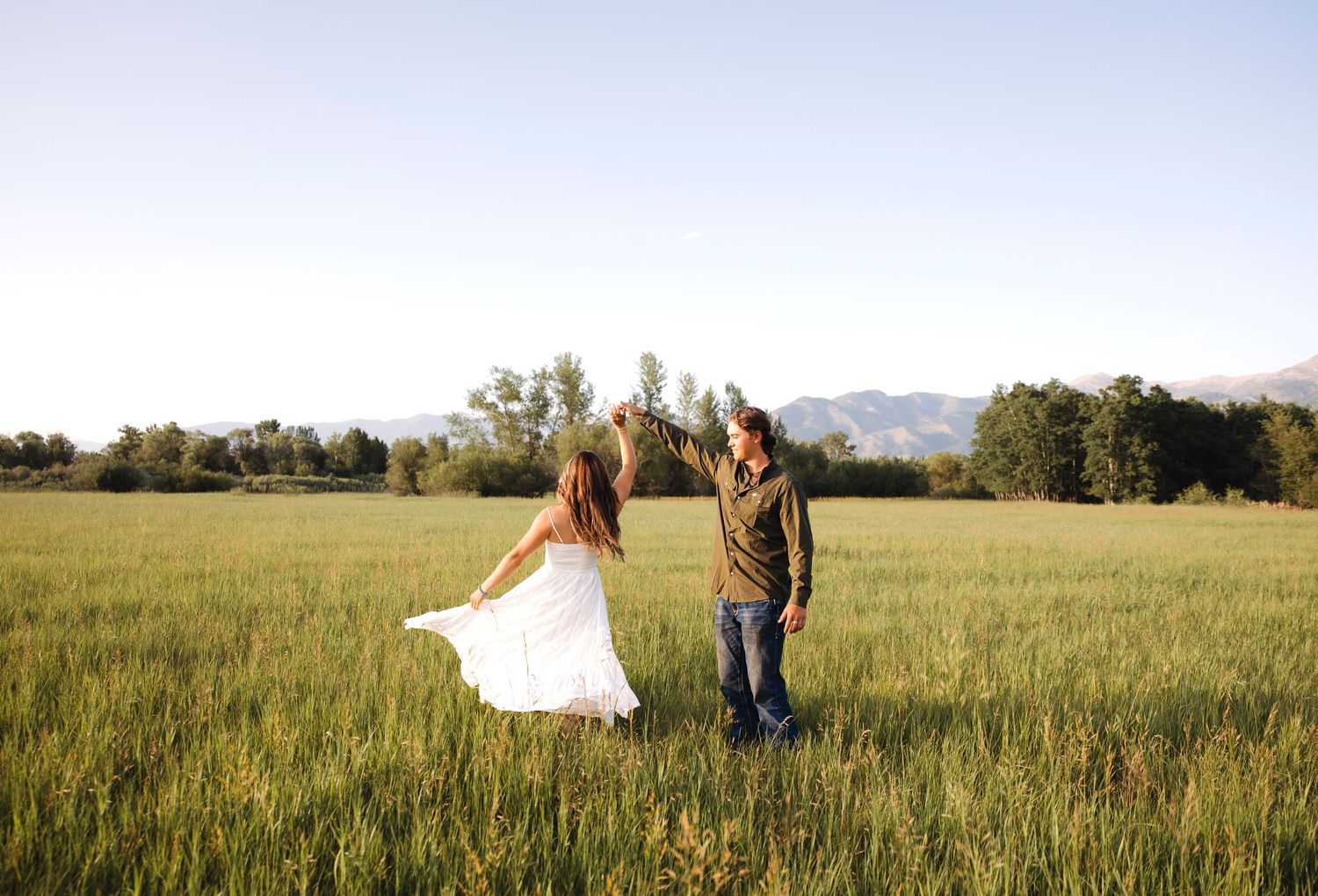 A couple walks hand in hand through a sunlit meadow with mountains in the background.