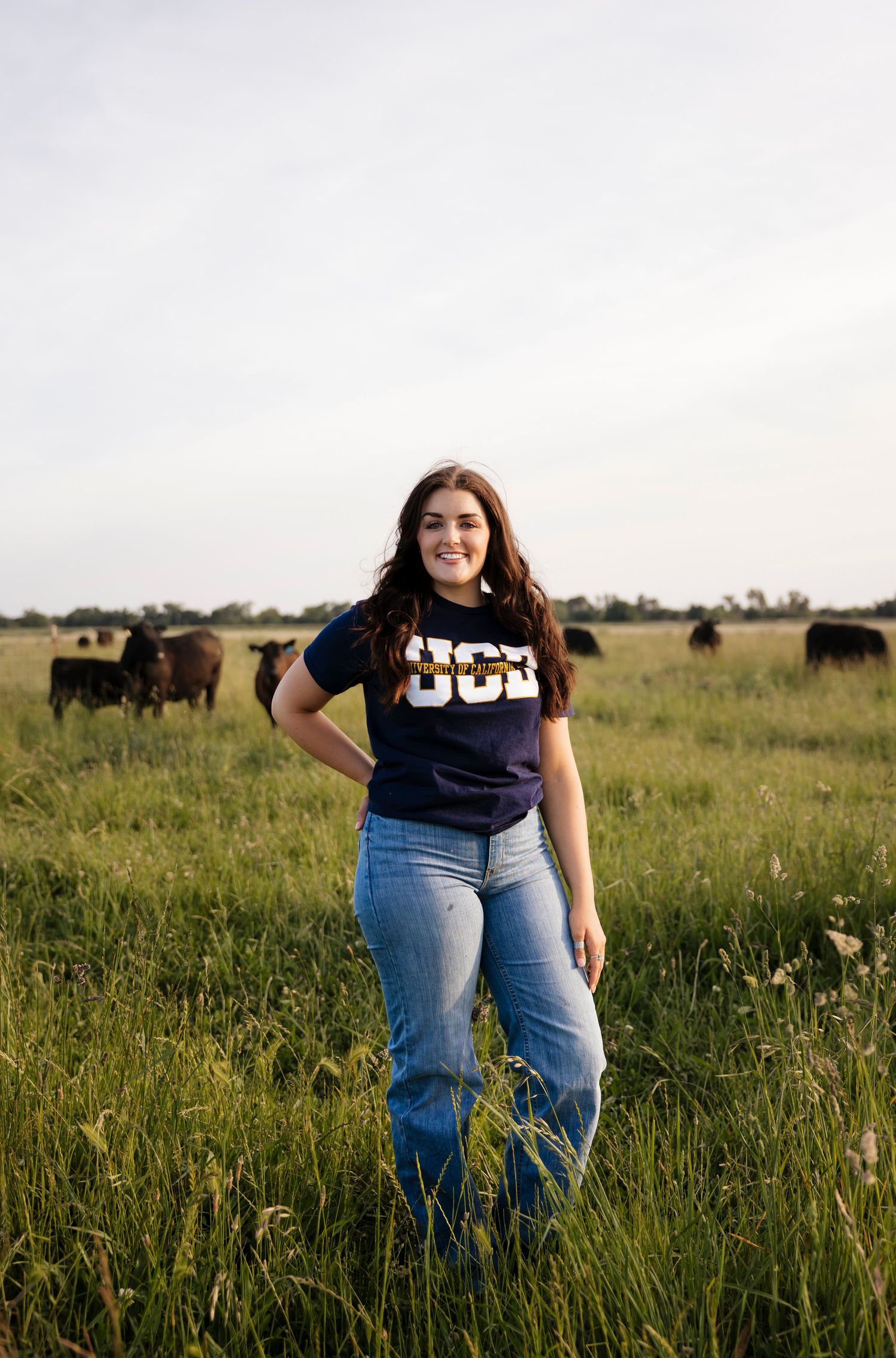 Graduate in UCM shirt poses in field with grazing cattle in background.