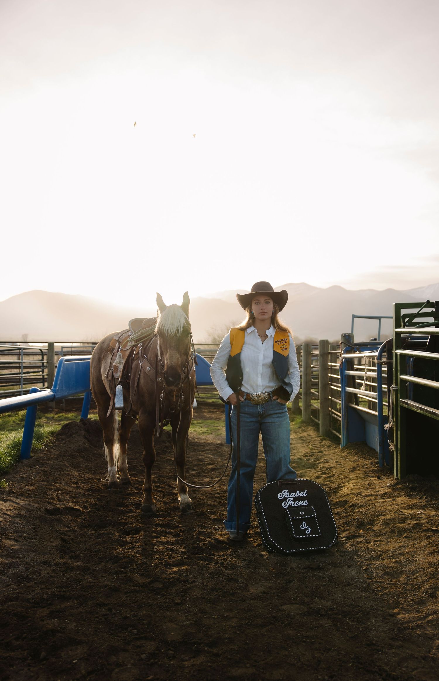 A cowgirl in a black hat and yellow vest stands with her horse in a rodeo arena at golden hour.