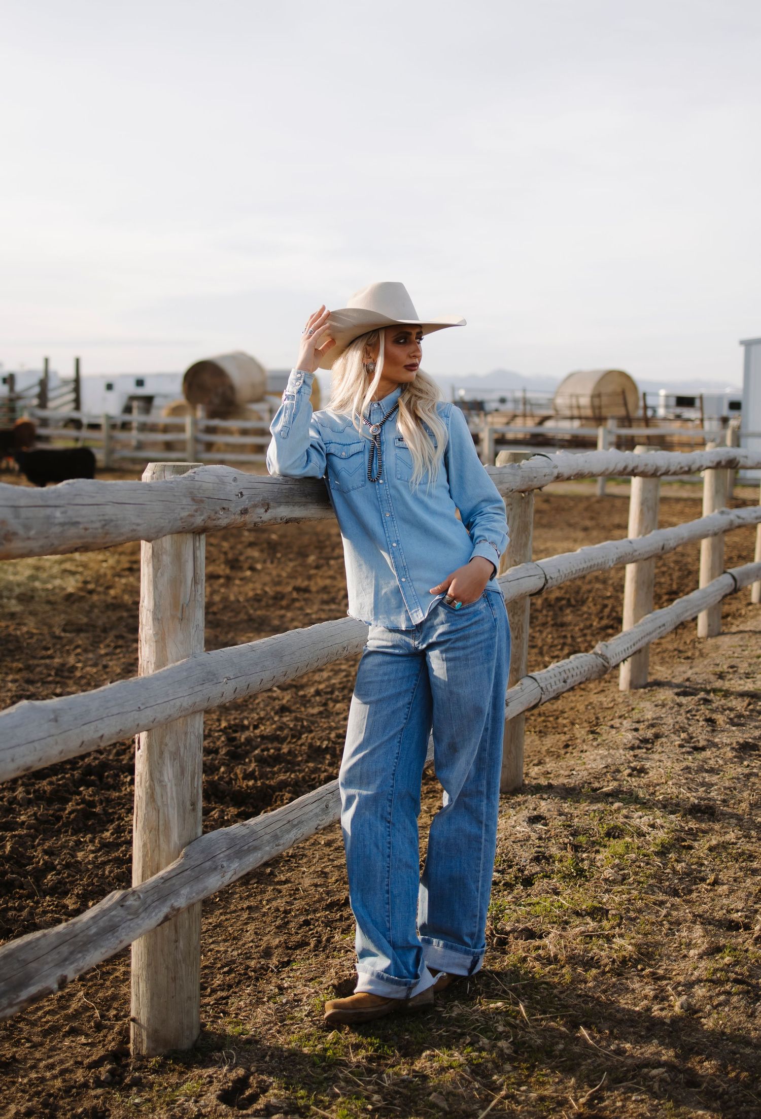 Blonde woman in cowboy hat, blue denim shirt, and jeans leaning on a wooden ranch fence outdoors.