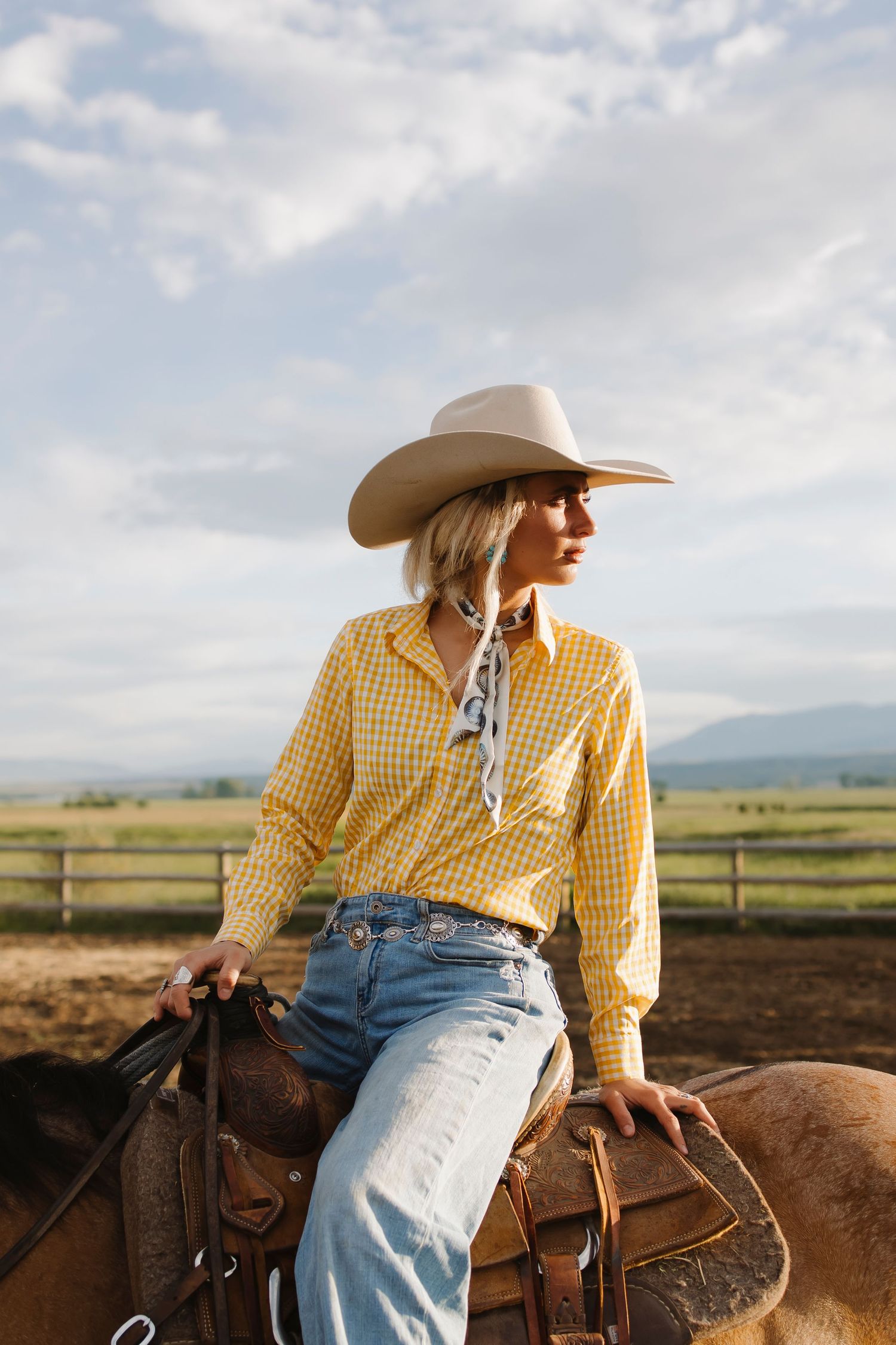 A cowgirl in a yellow plaid shirt, jeans, and white cowboy hat sits on a horse at a ranch with mountain views.
