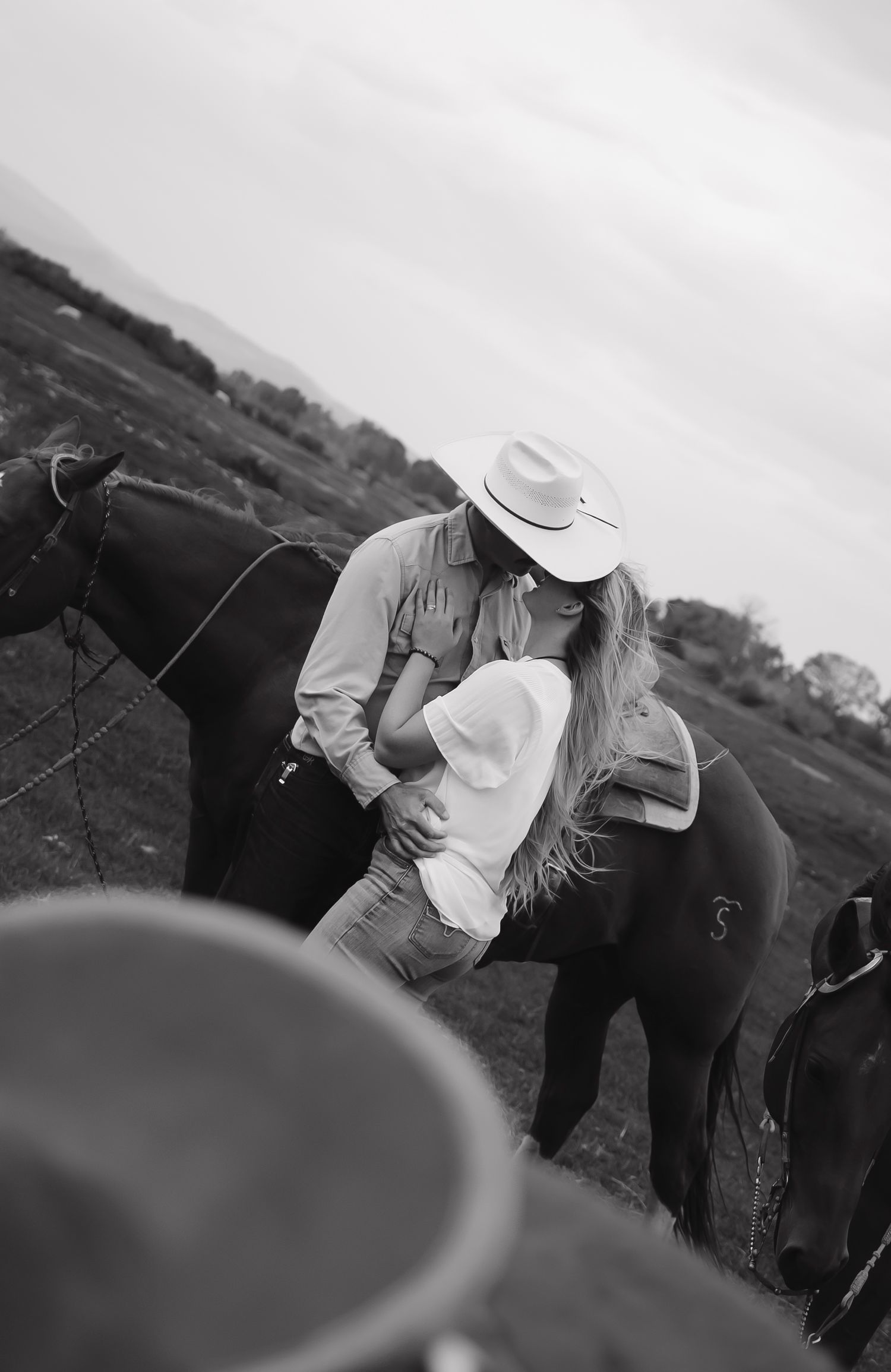 A romantic black and white photo of a couple embracing near horses.