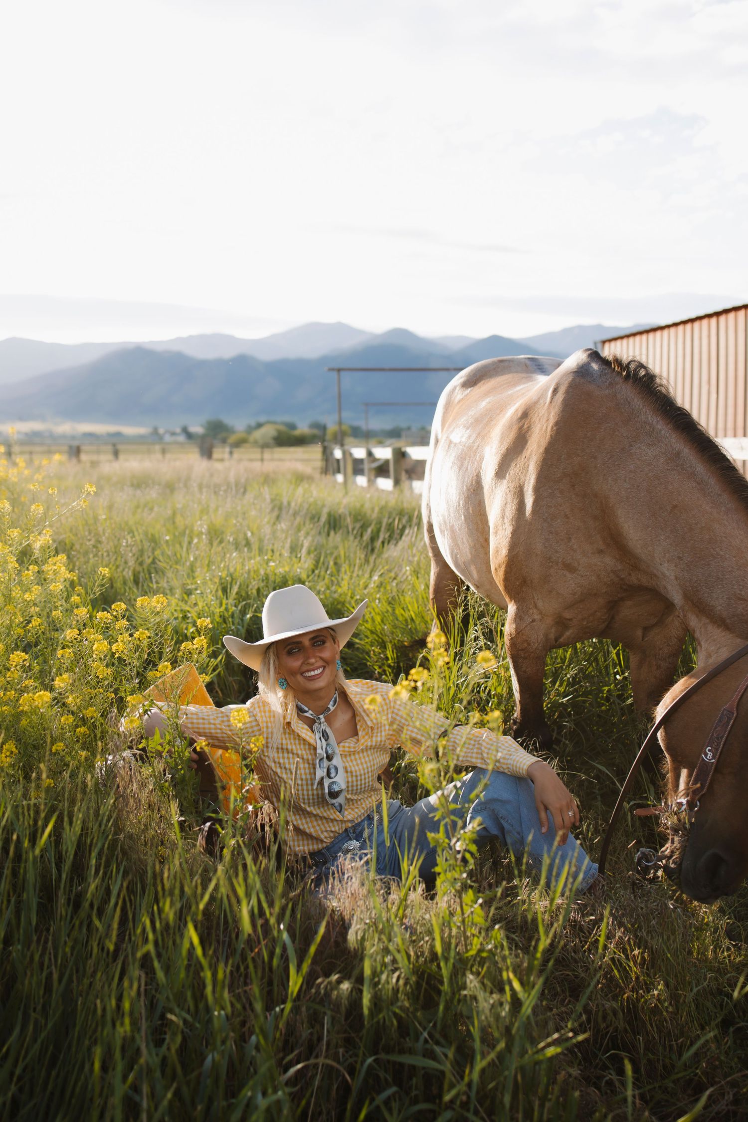 Smiling cowgirl in white hat sits in green field with yellow flowers beside a grazing horse, mountains in background.