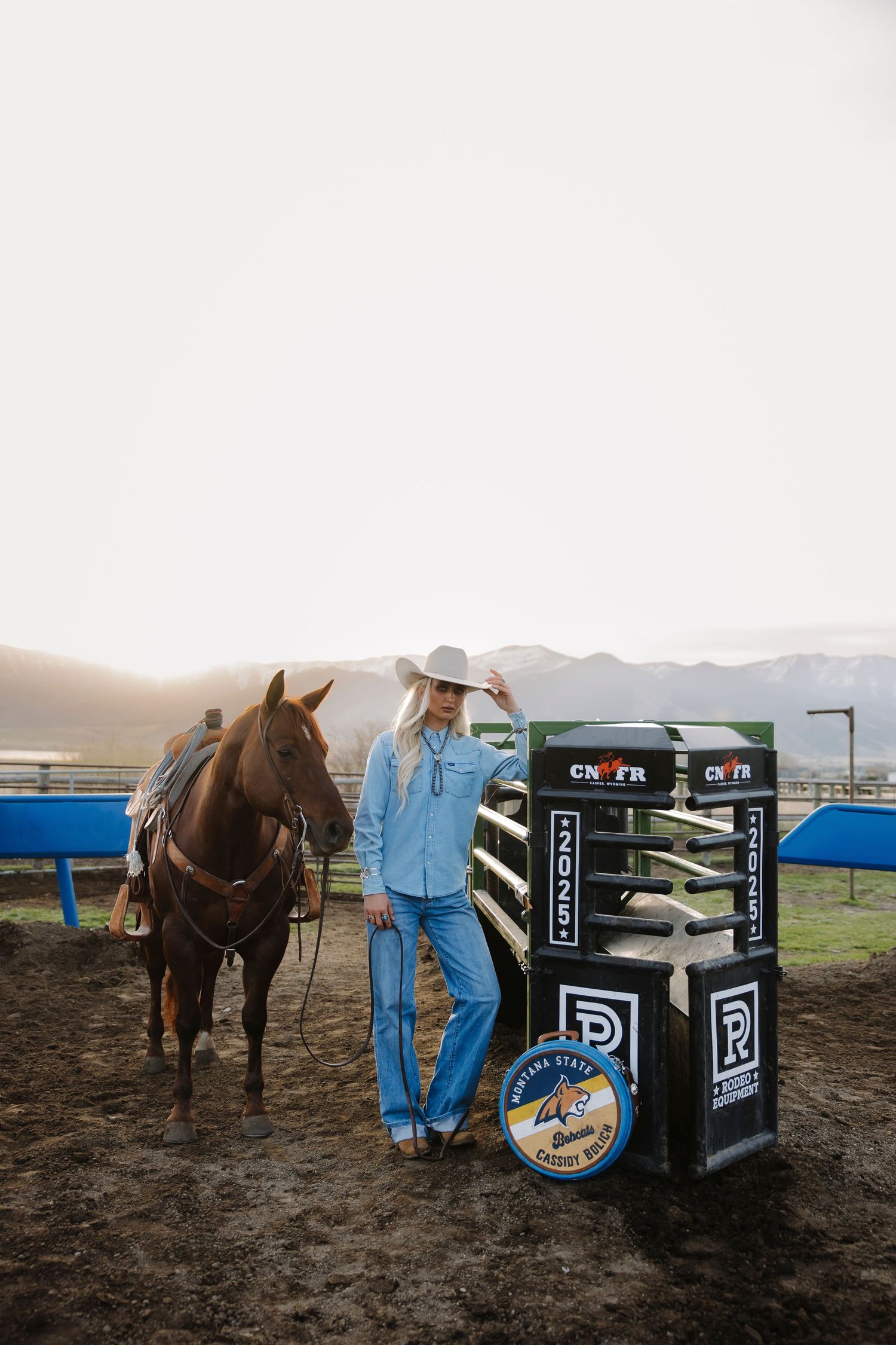 Cowgirl in white hat and blue denim stands beside a chestnut horse near CNFR rodeo signage at golden hour.