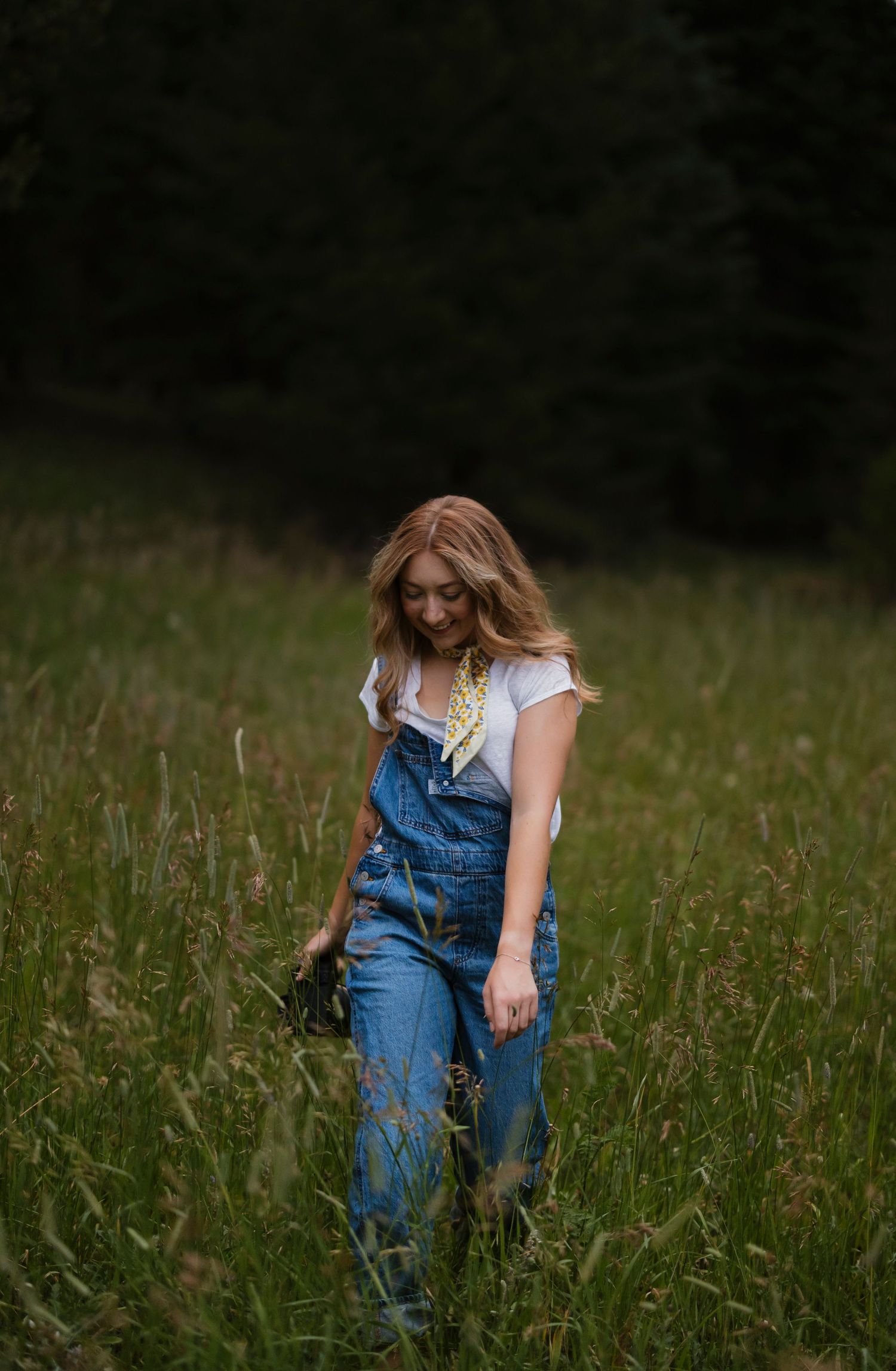 Young woman in denim overalls and white tee walks through tall green meadow grass with dark trees behind her.