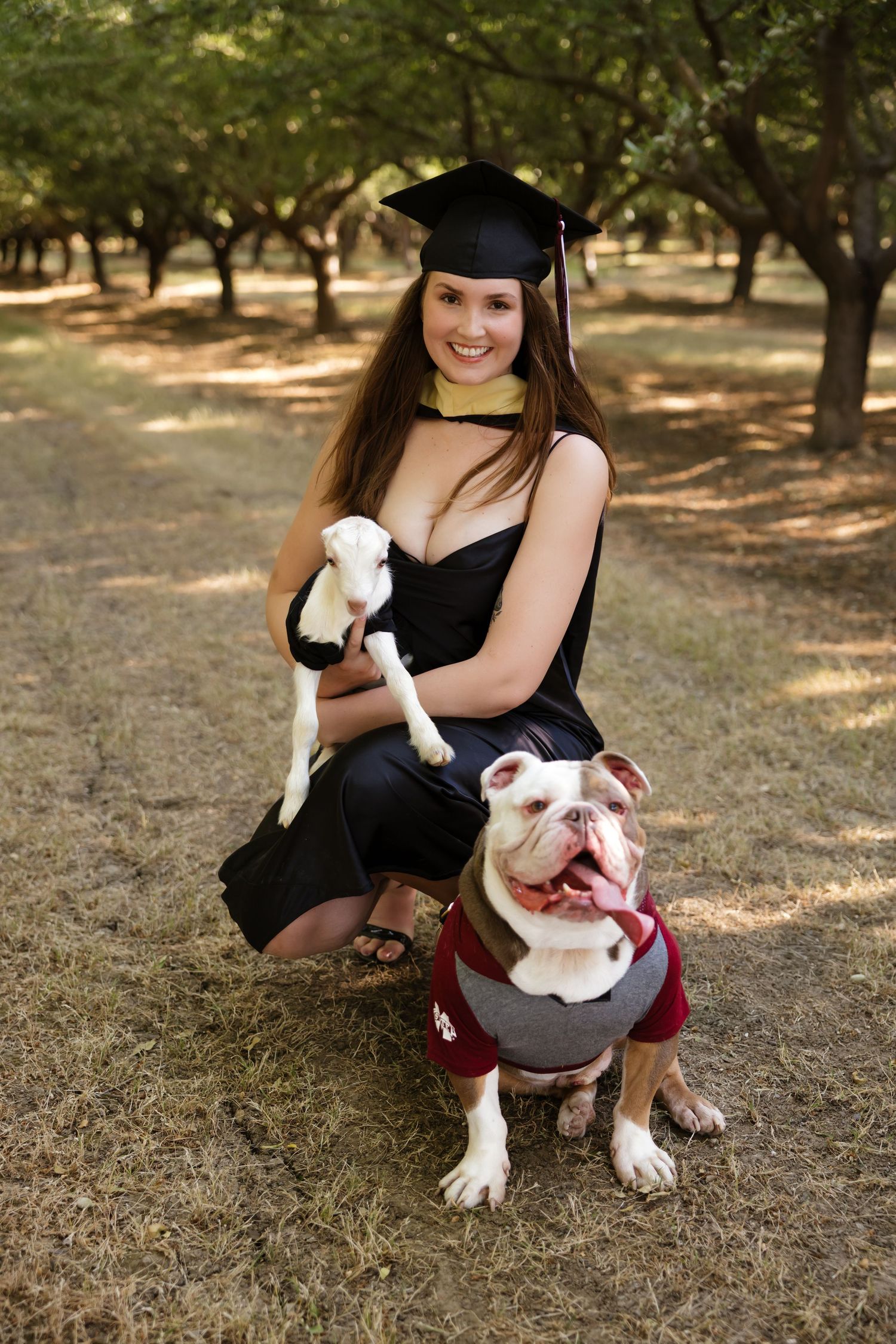 Graduate in cap and gown kneels with bulldog wearing matching graduation attire.