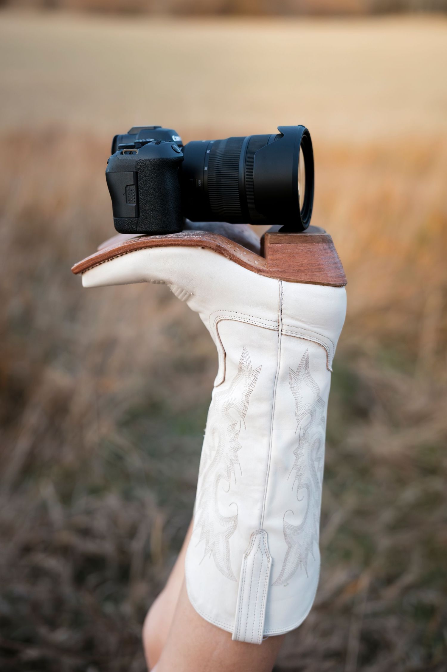 A black DSLR camera balanced on top of a white cowboy boot held up against a blurred field background.