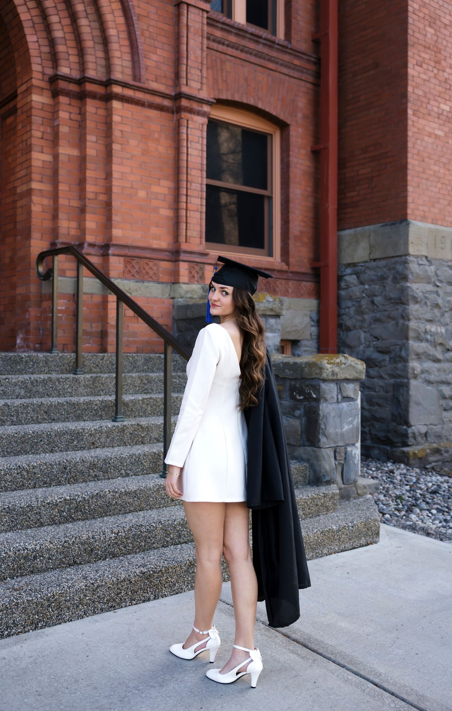 Graduate in white dress and black cap poses on brick building steps.