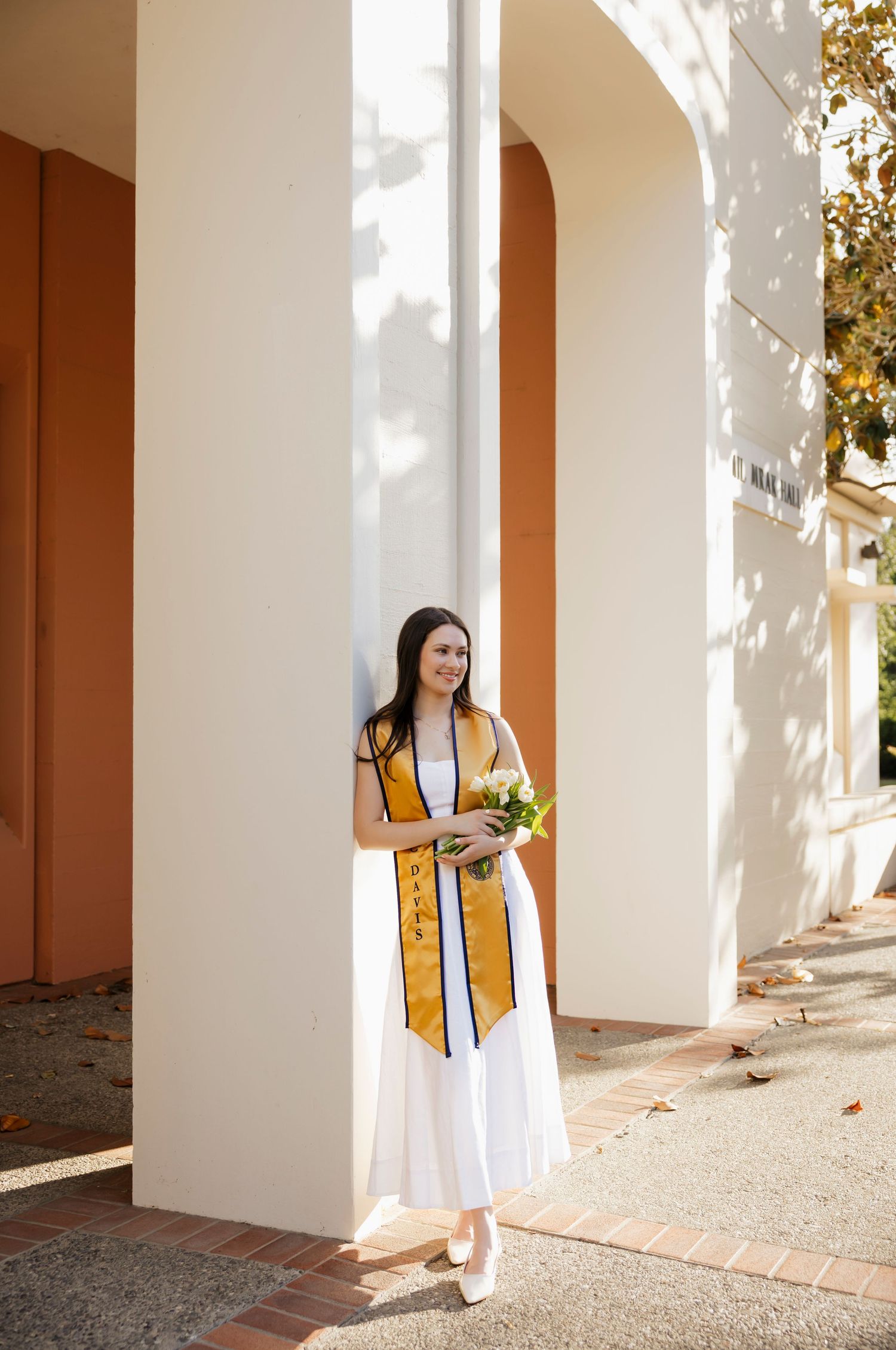 Person in white dress stands between architectural columns with dappled sunlight.