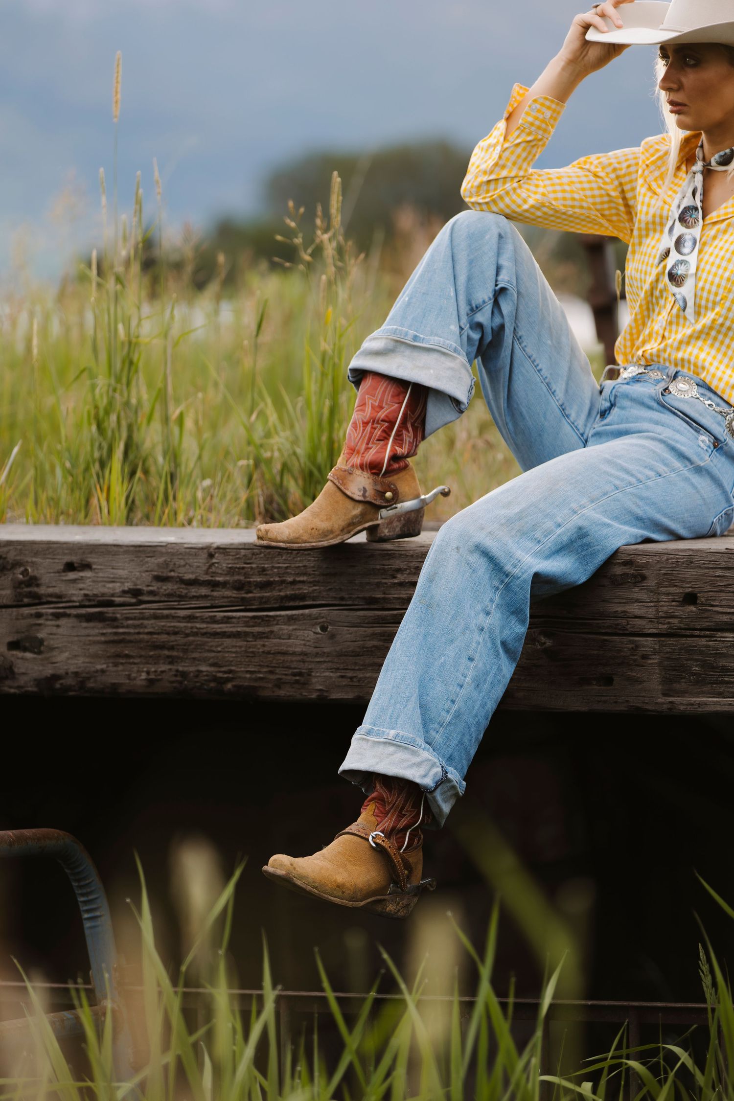 A cowgirl in jeans, yellow plaid shirt, and cowboy boots with spurs sits on a wooden fence rail in a grassy field.