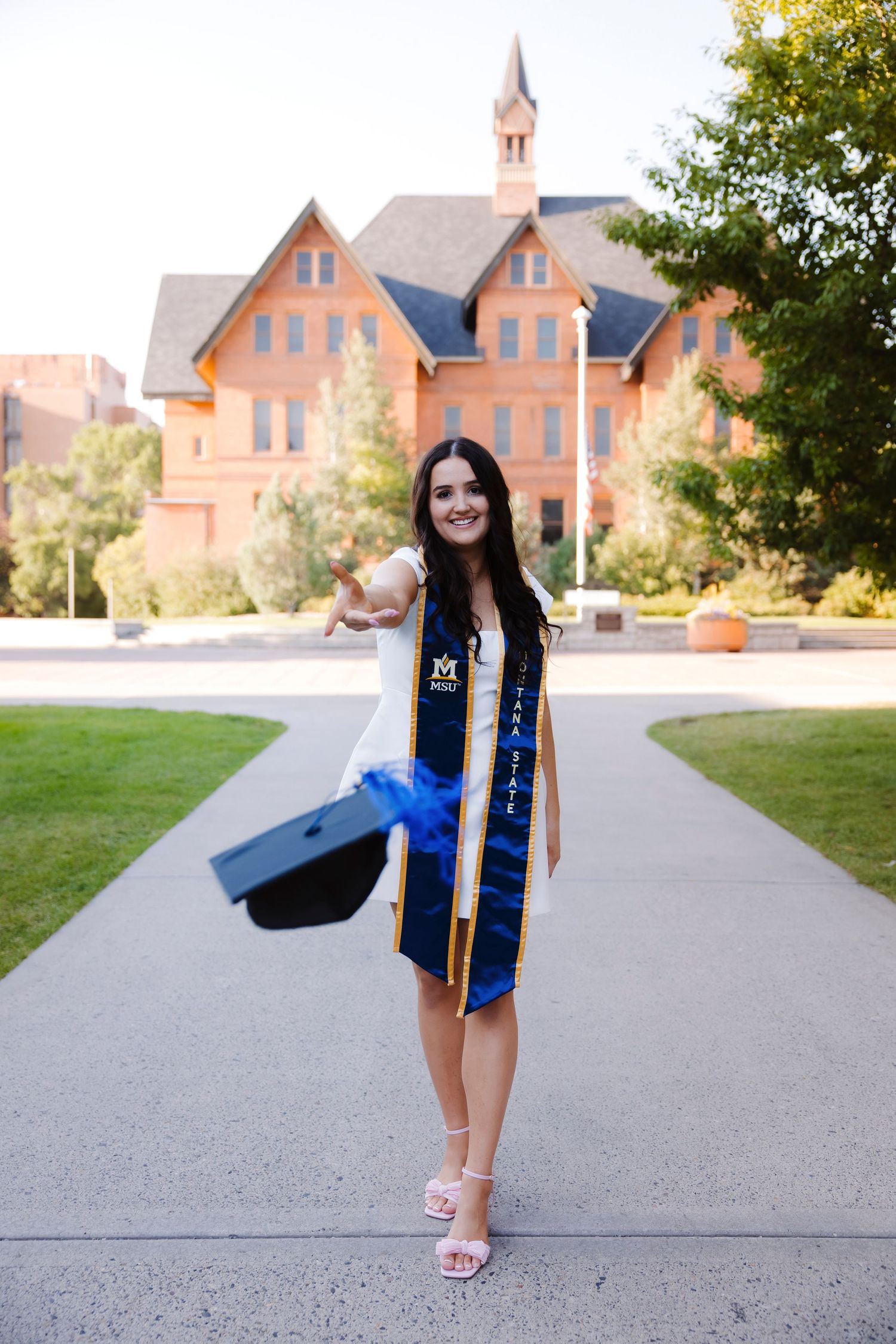 Graduate celebrates on campus path with historic brick buildings in background.