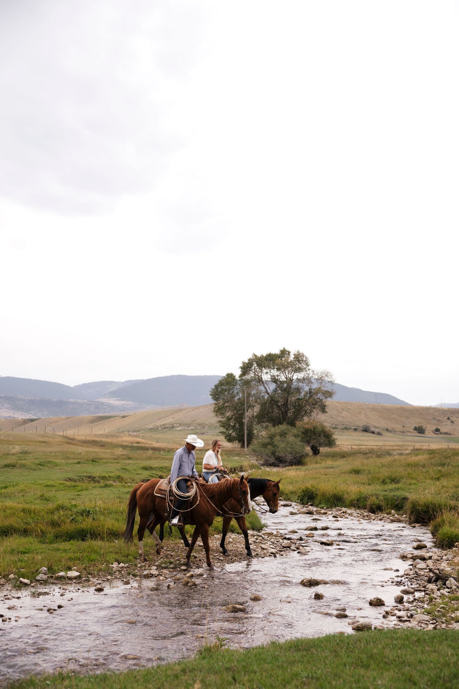 Horses and riders cross a rocky stream in a picturesque mountain valley setting.
