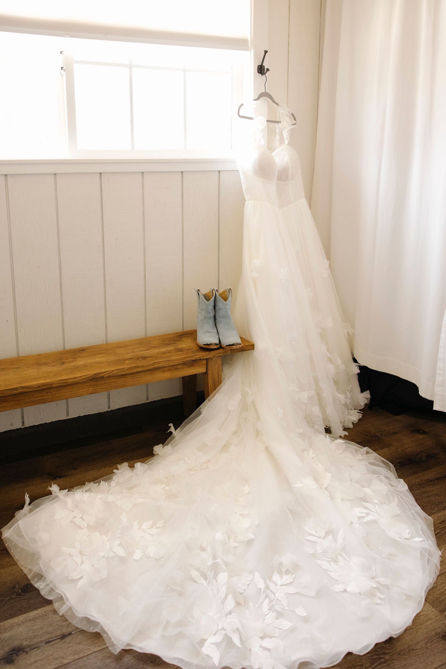 A stunning white wedding gown with floral appliqués and long train hangs near a window, with blue cowboy boots on a wooden bench.