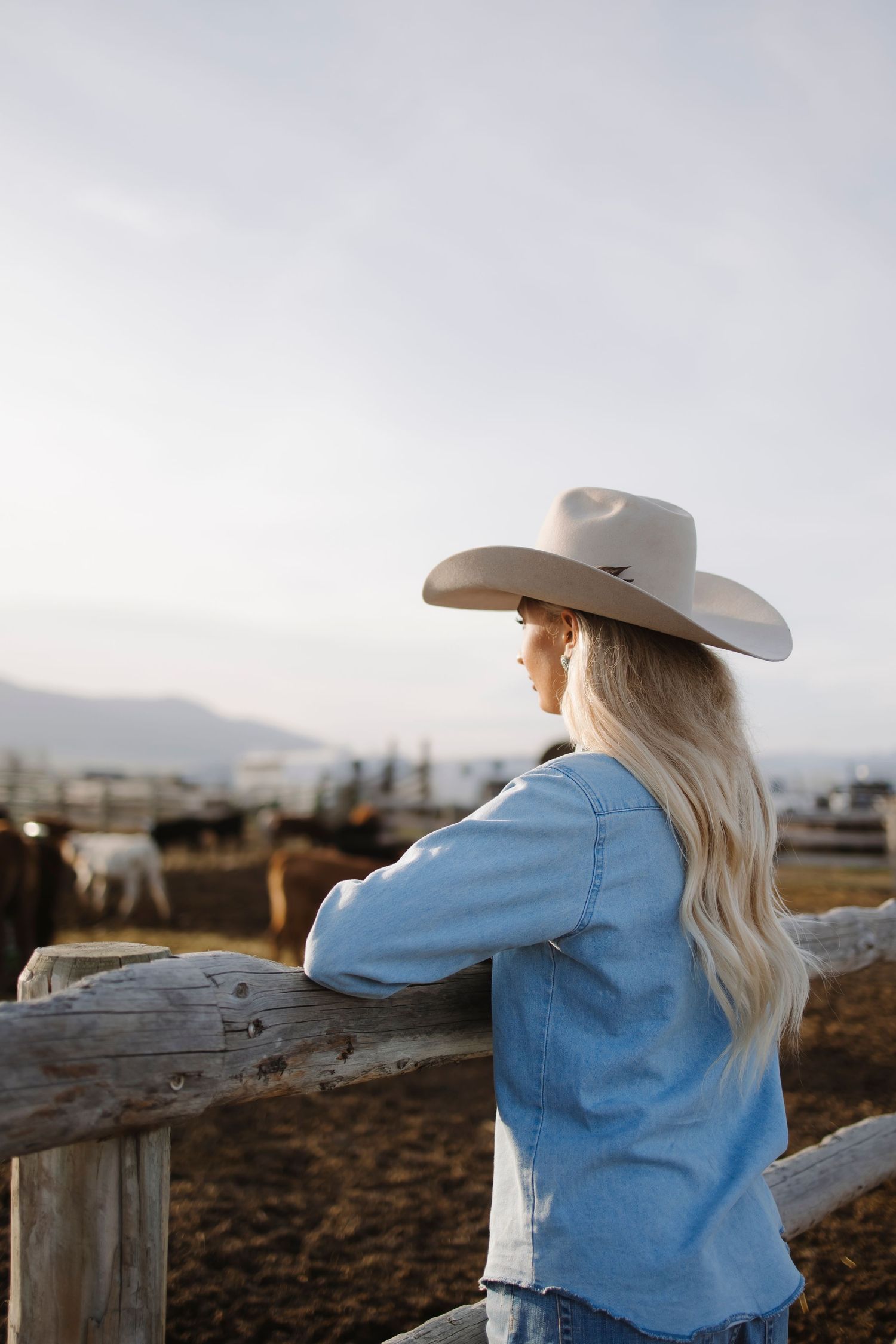 Blonde woman in cowboy hat and denim shirt leaning on wooden fence overlooking cattle ranch at sunset.