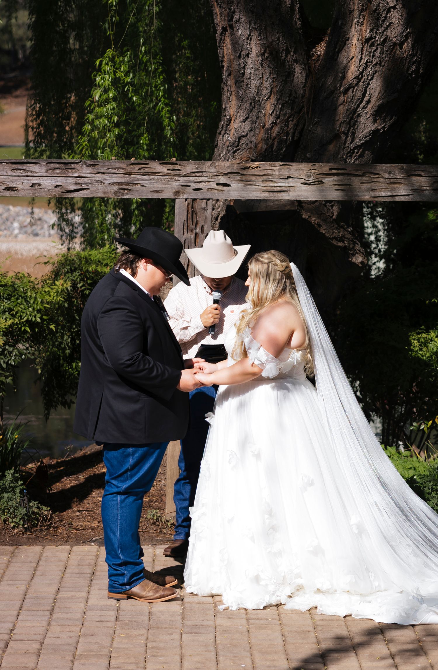 Western wedding ceremony taking place on a brick pathway beneath a large willow tree.
