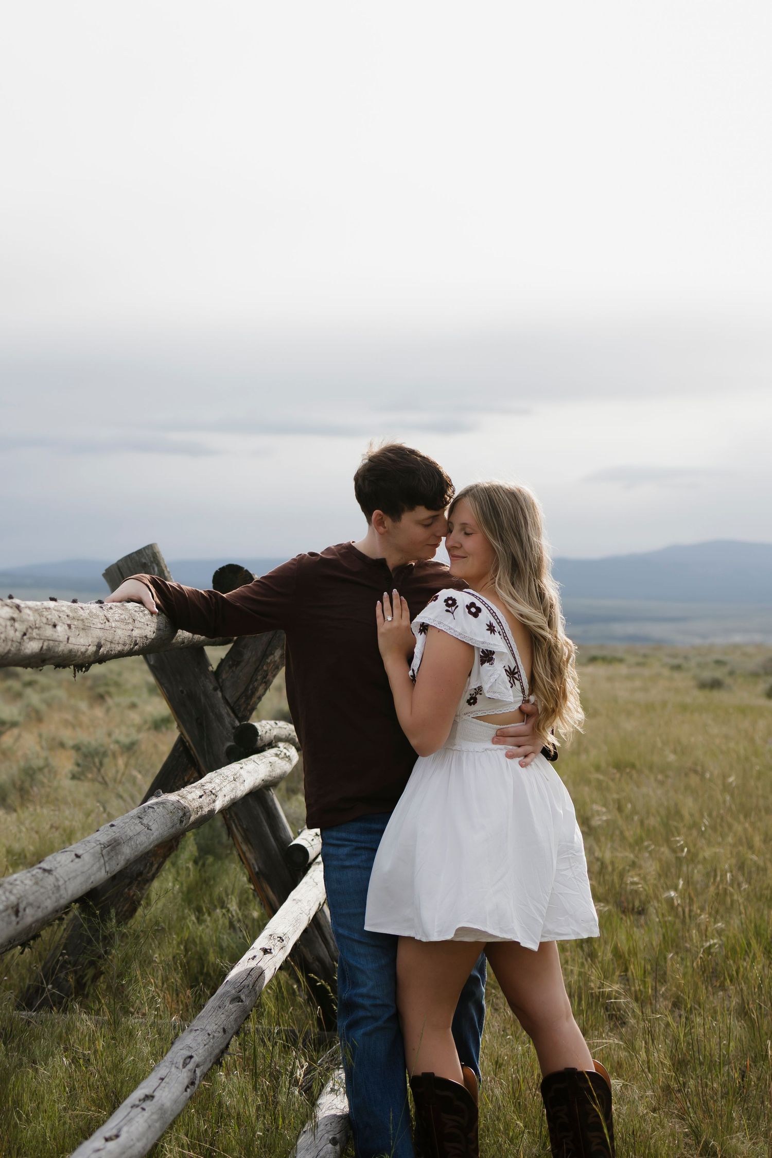 A couple shares a romantic moment by a rustic wooden fence in a grassy field, she in a white dress and boots.