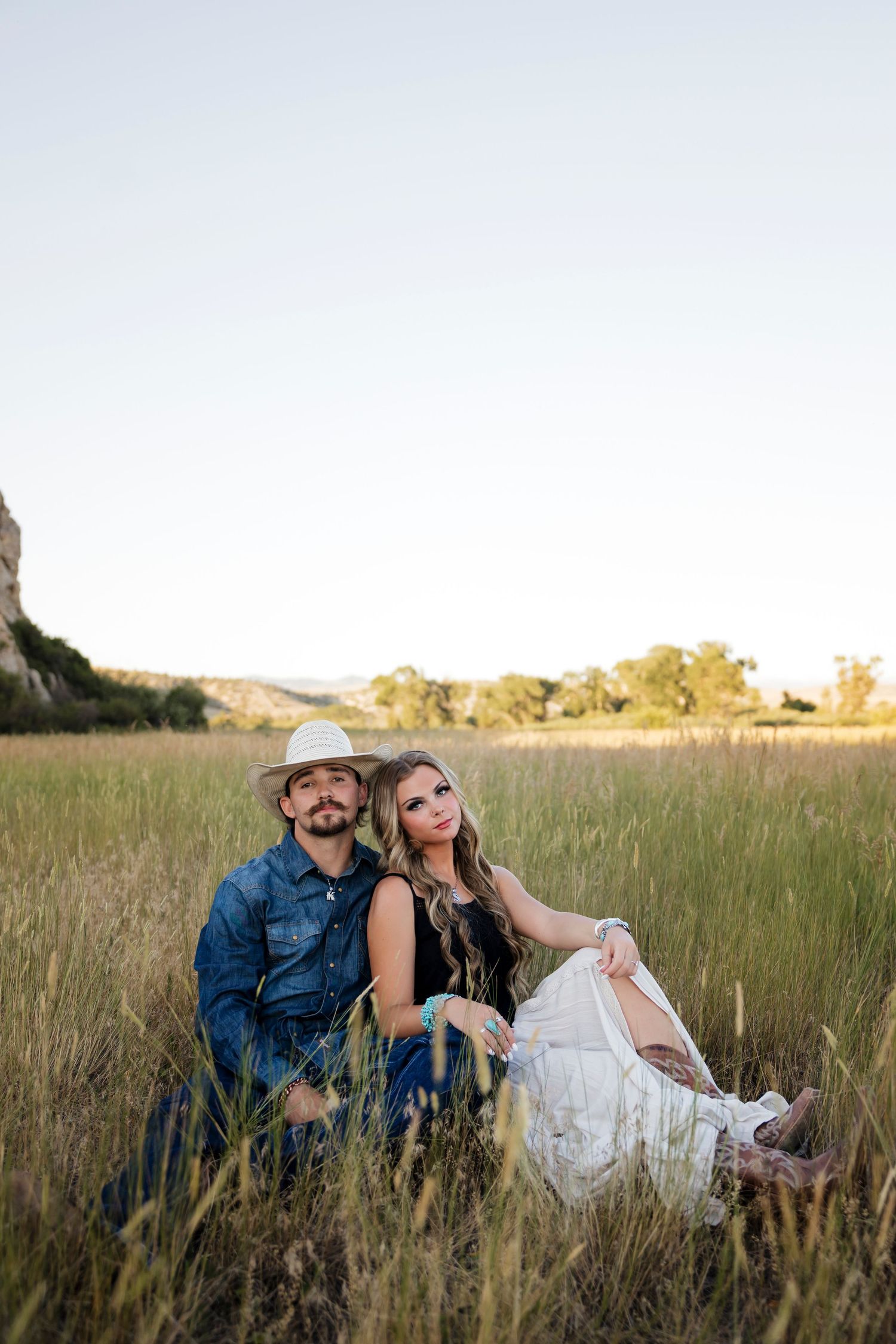 A romantic couple sits together in tall grass at sunset near rocky cliffs.