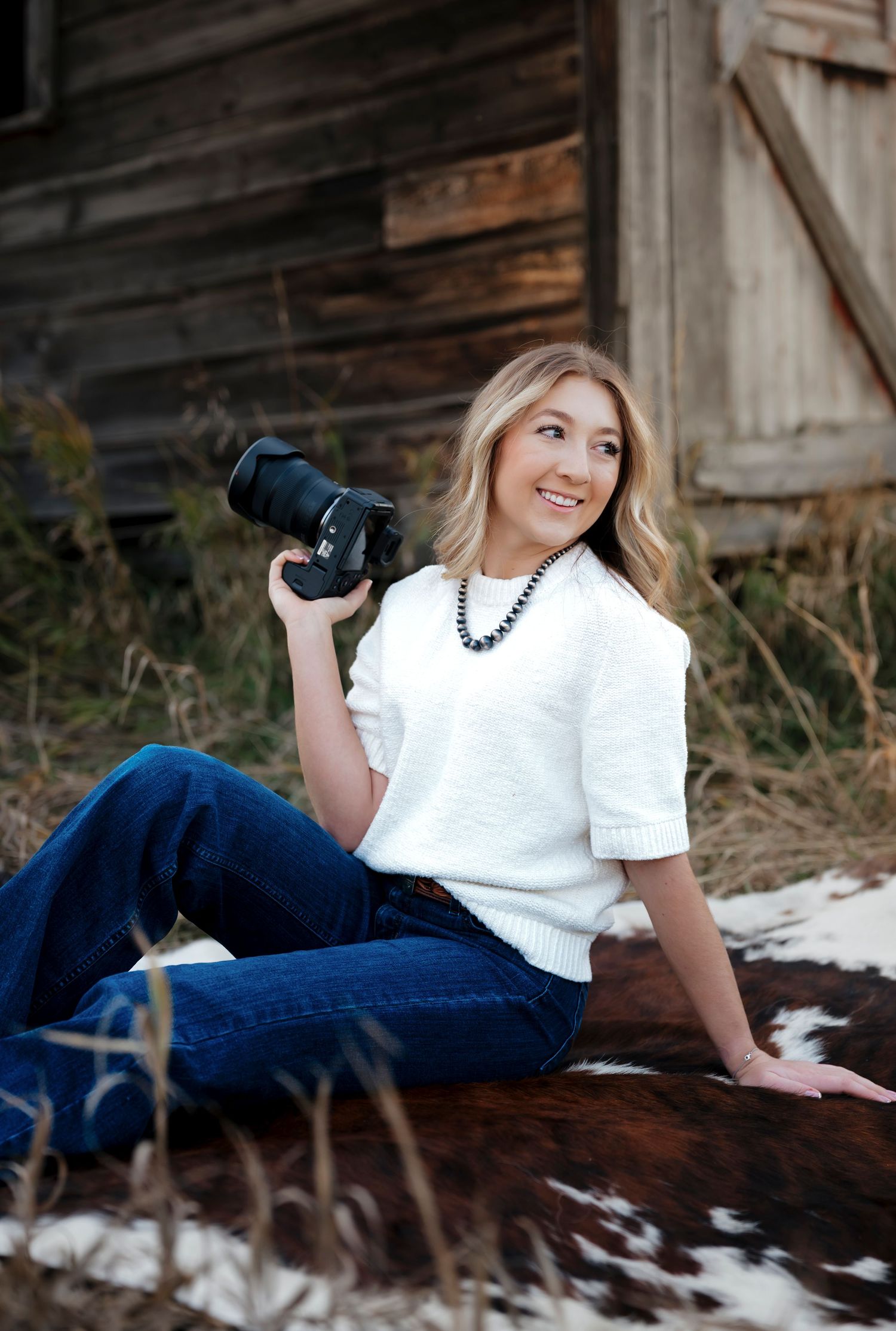 Smiling blonde woman holding a DSLR camera, wearing white sweater and jeans, sitting outdoors near a rustic wooden barn.