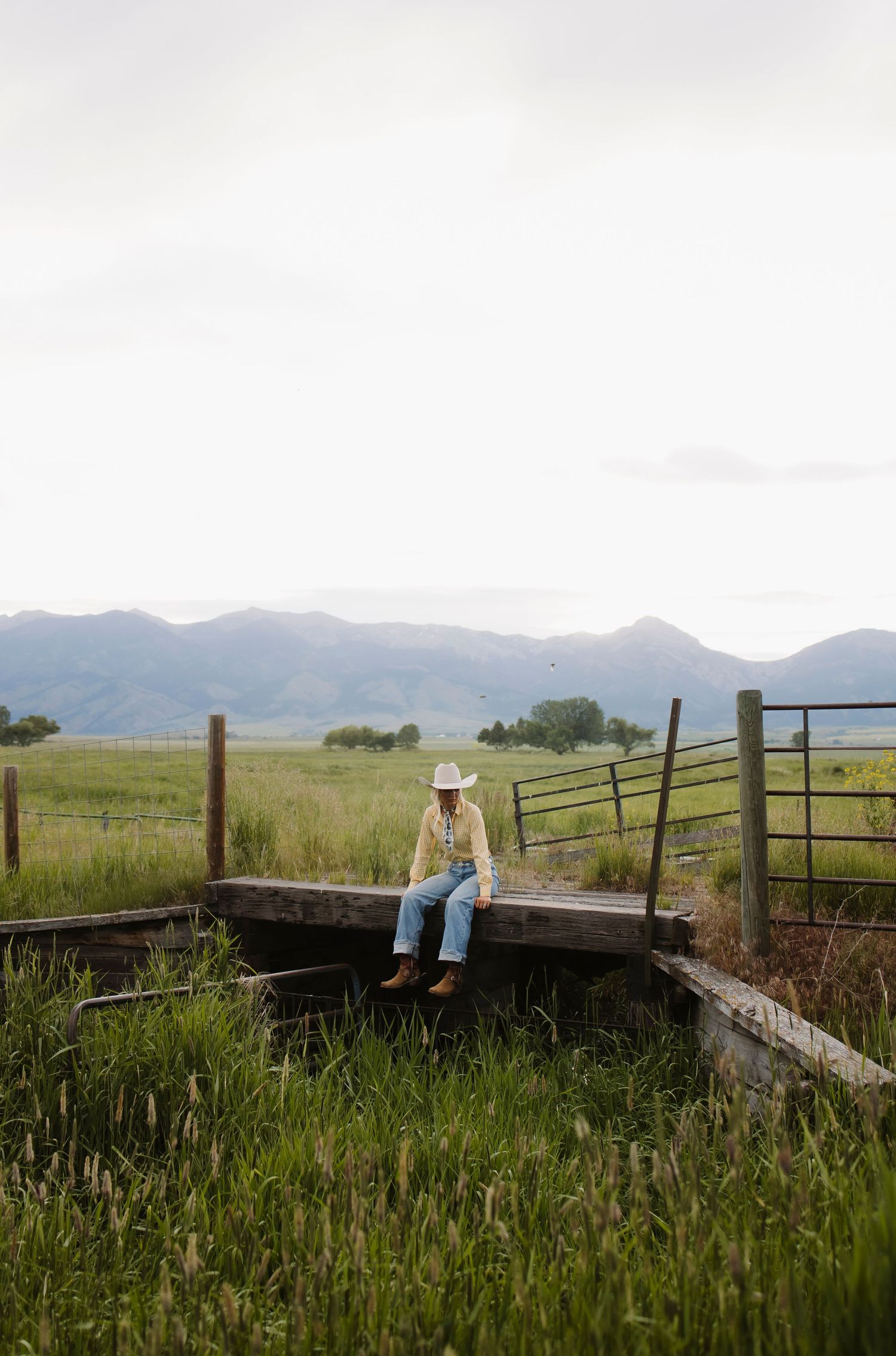 A cowboy in a white hat and jeans sits on a wooden fence in a vast green meadow with mountains in the background.