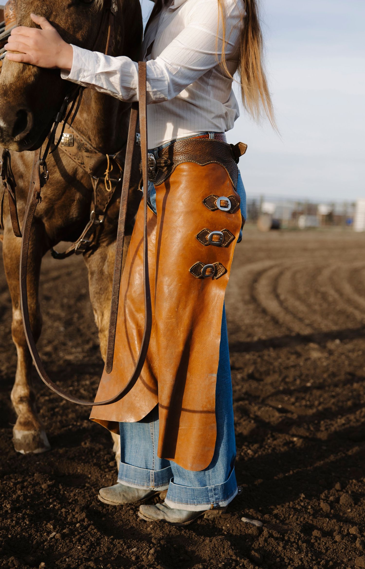 Cowgirl in leather chaps and jeans holds horse reins on a dirt arena, showcasing western riding apparel.