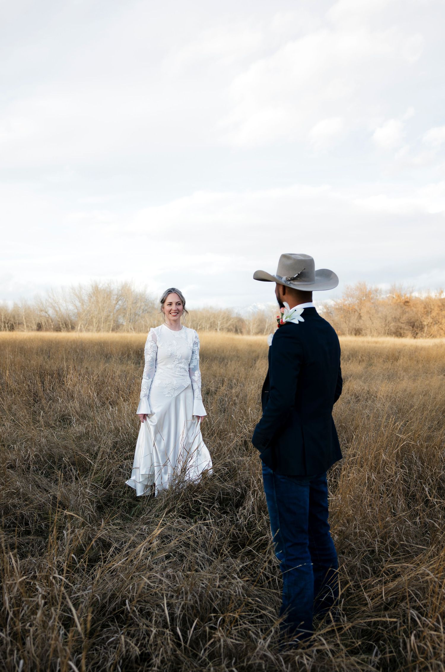 Couple in wedding attire standing apart in tall grass field during photoshoot.