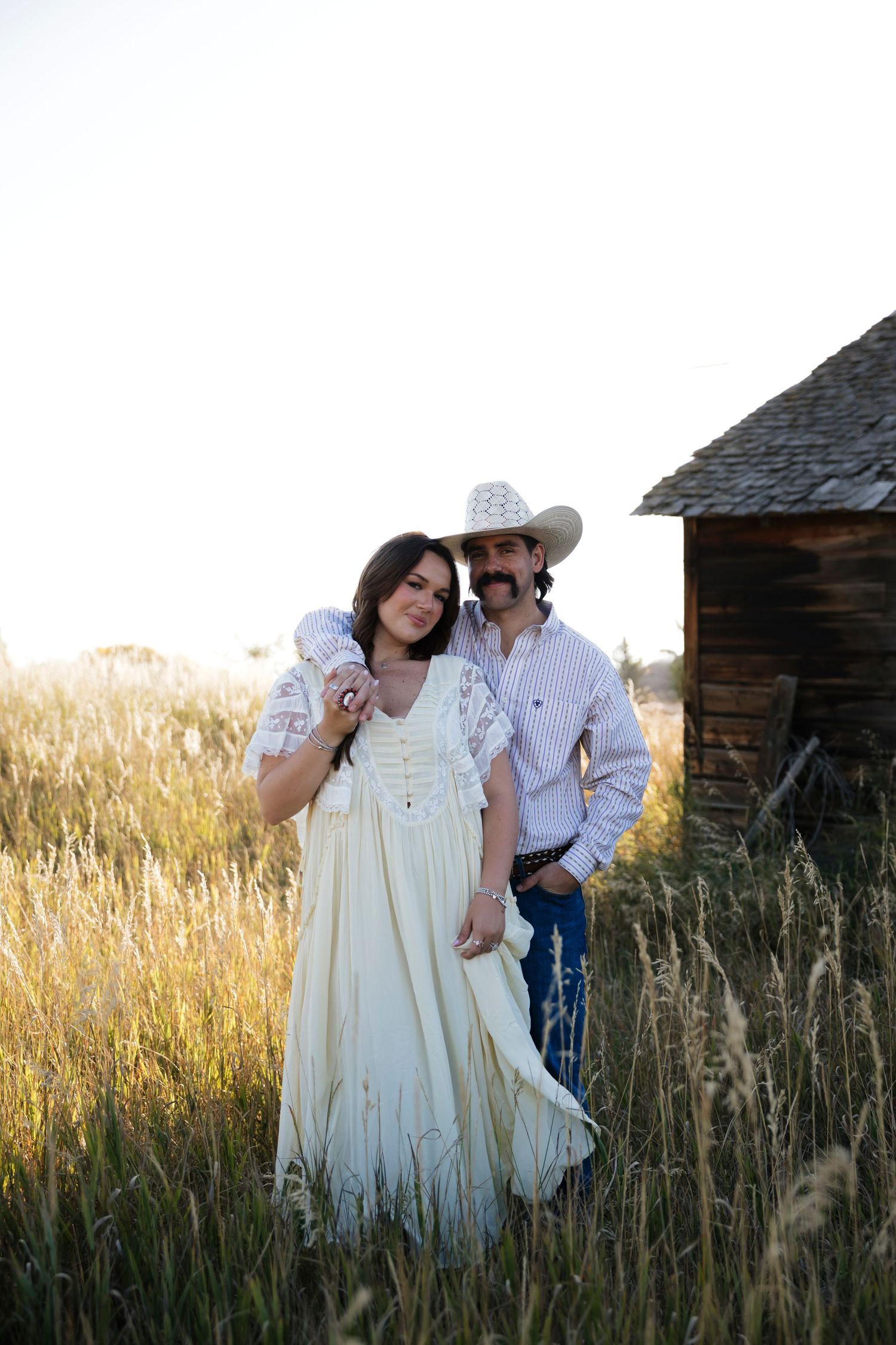 Couple embraces near rustic barn surrounded by golden prairie grass at sunset.