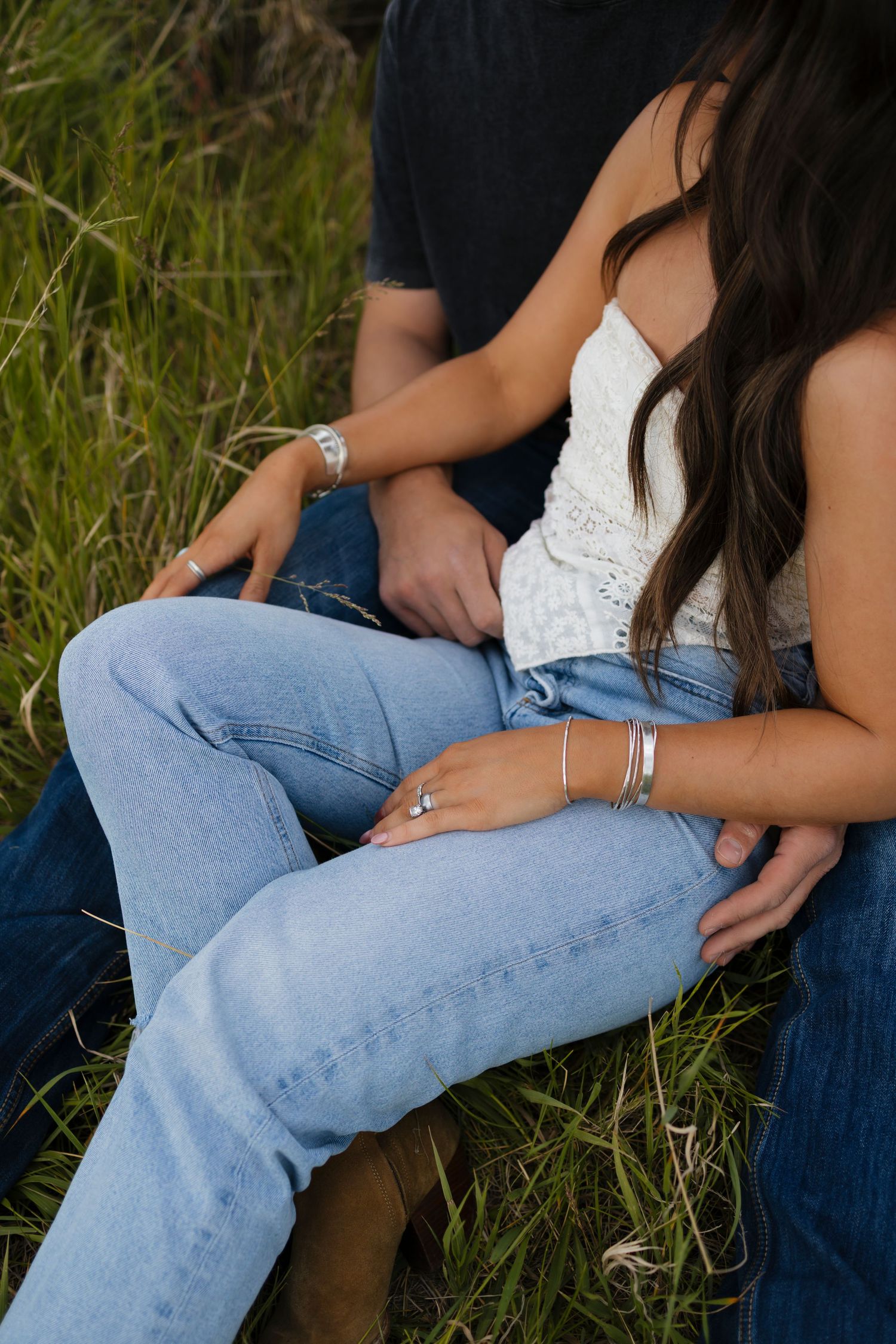 Couple sitting together in green grass, woman wearing white lace top, blue jeans, silver rings and bracelets.