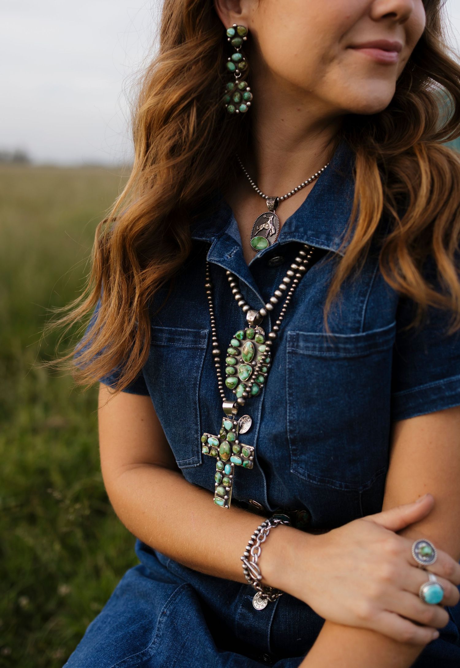Woman in denim jacket wearing layered turquoise necklaces, cross pendant, matching earrings, silver bracelet, and turquoise ring.