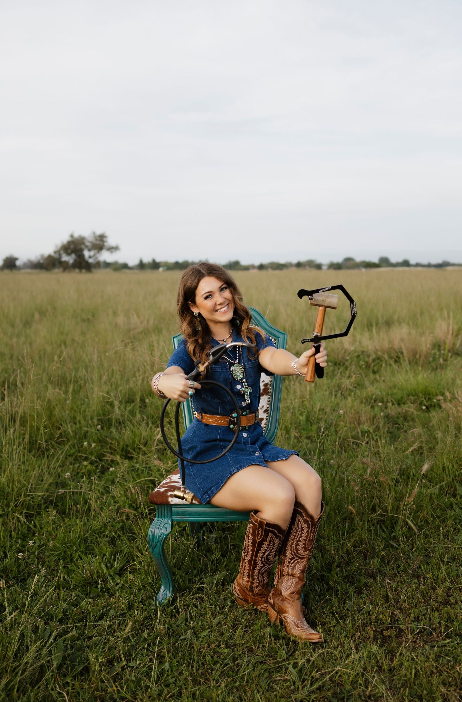 Smiling woman in blue dress and cowboy boots sits on green chair in grassy field holding a slingshot.