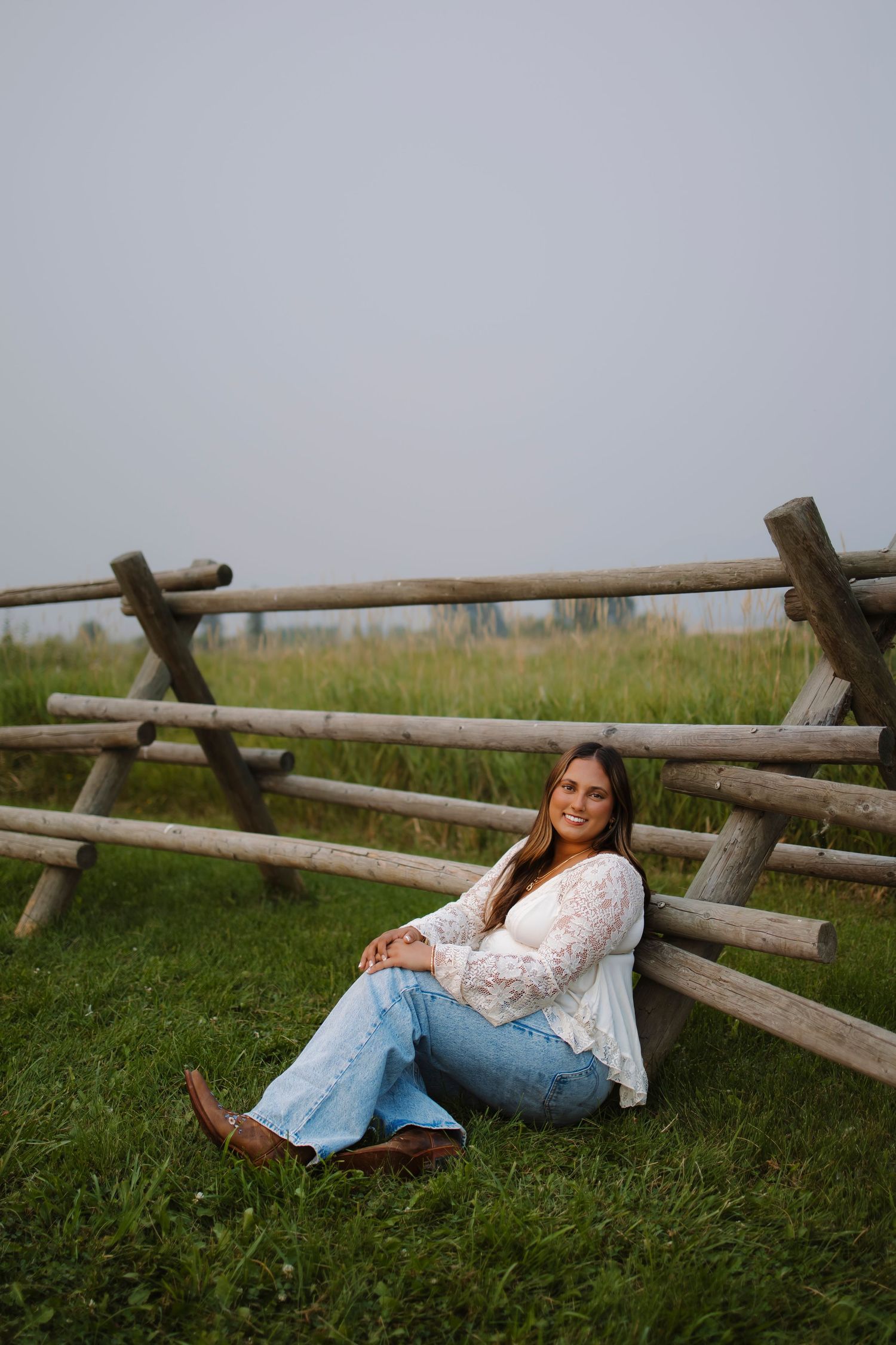 Young woman in white lace top, jeans, and cowboy boots sitting against a rustic wooden fence on green grass.
