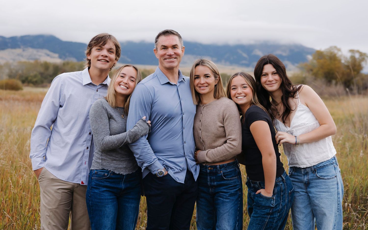 Family of six smiling together in a scenic mountain meadow, wearing casual outfits for an outdoor portrait.
