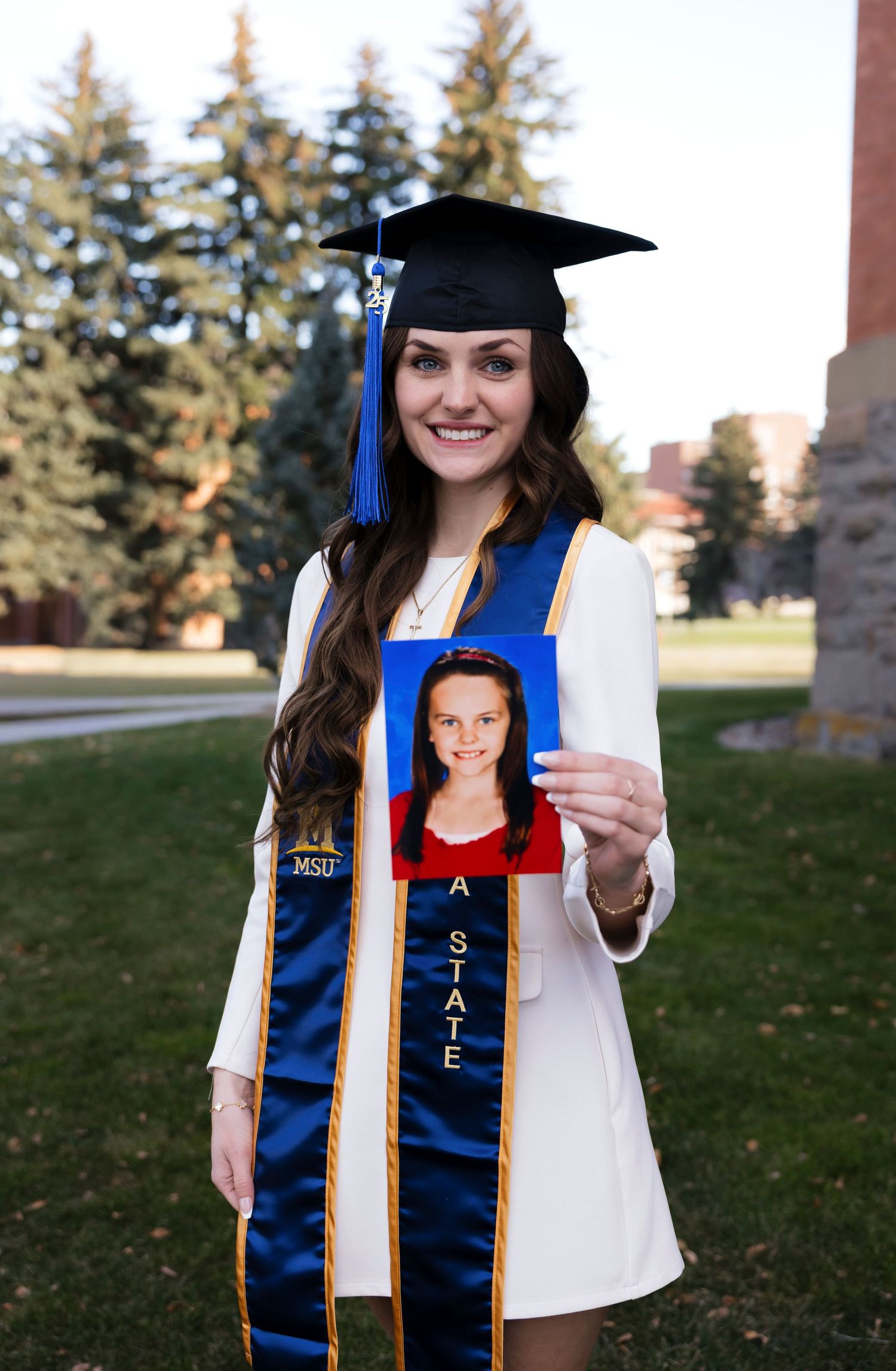 Graduate wearing blue and gold graduation stole and cap poses with diploma outdoors.