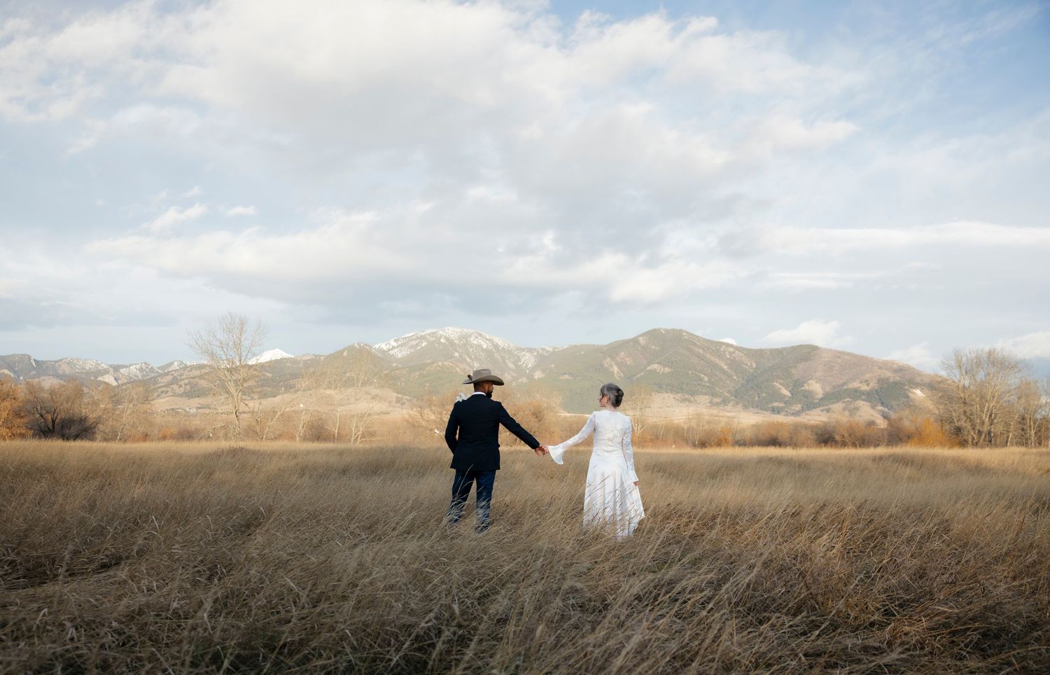 Couple in wedding attire holding hands in golden field with mountain backdrop under cloudy sky.