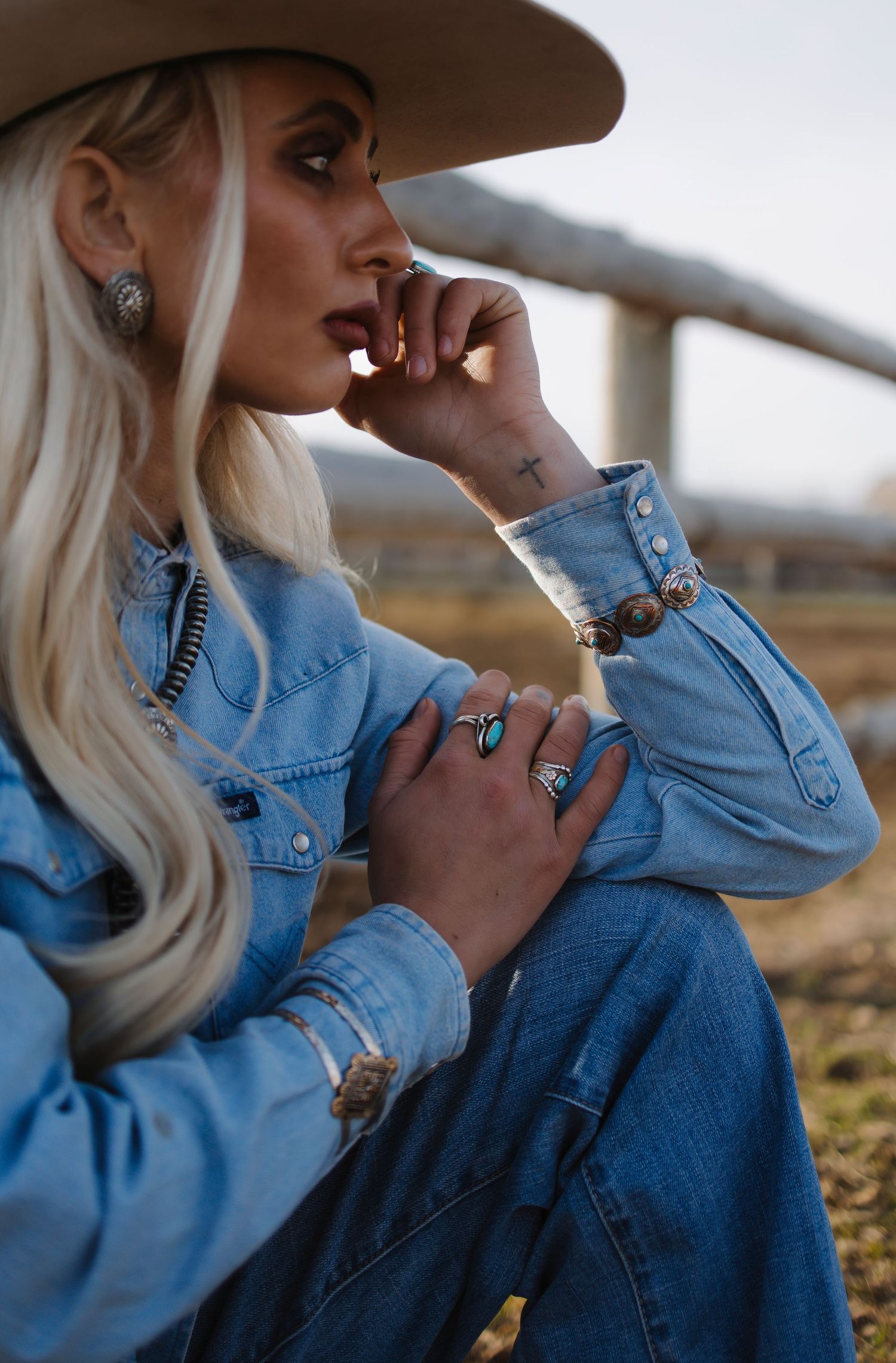 Blonde woman in denim shirt and wide-brim hat wearing turquoise and silver jewelry at an outdoor ranch setting.
