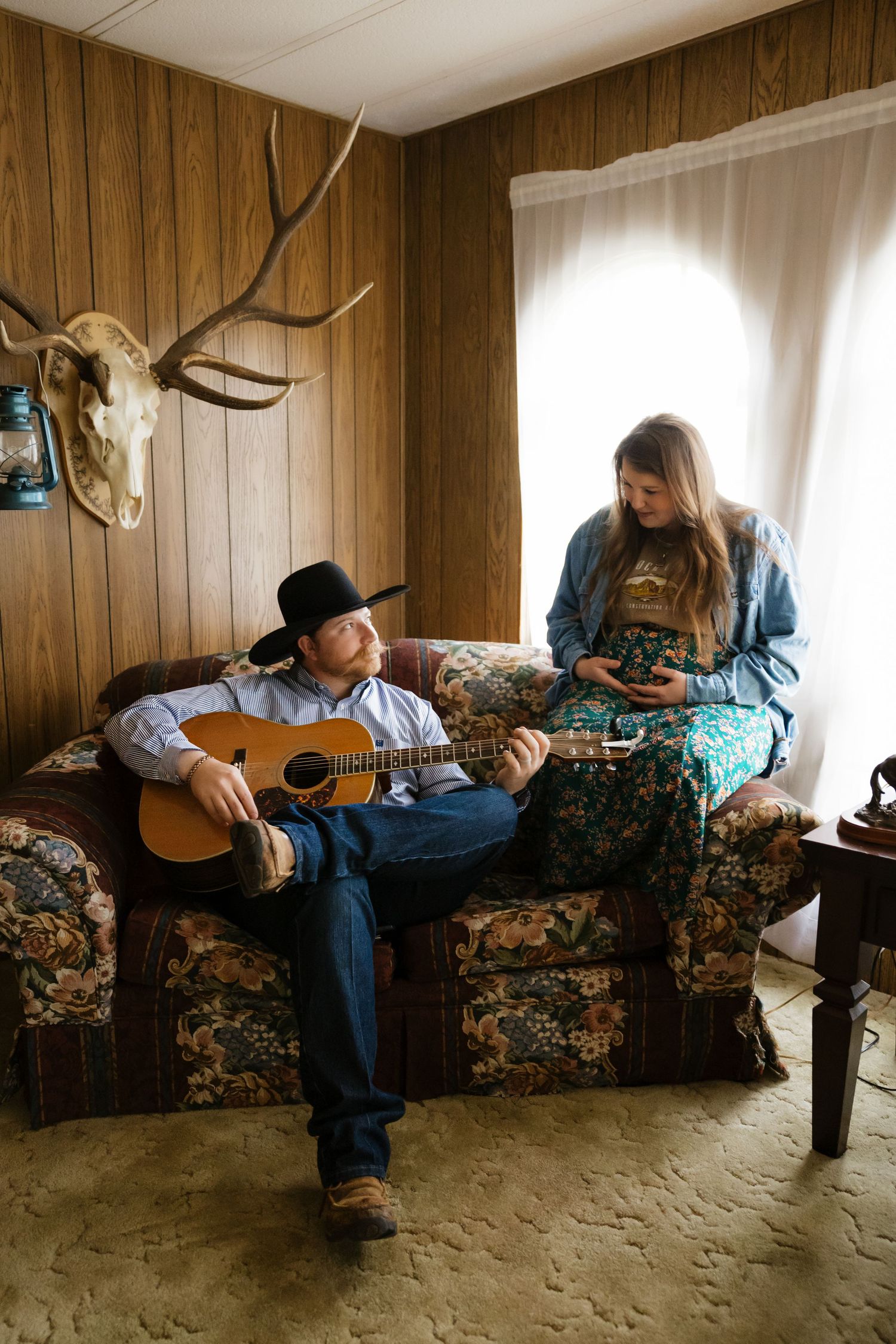 Musicians playing guitar and relaxing on floral couch beneath mounted deer antlers.