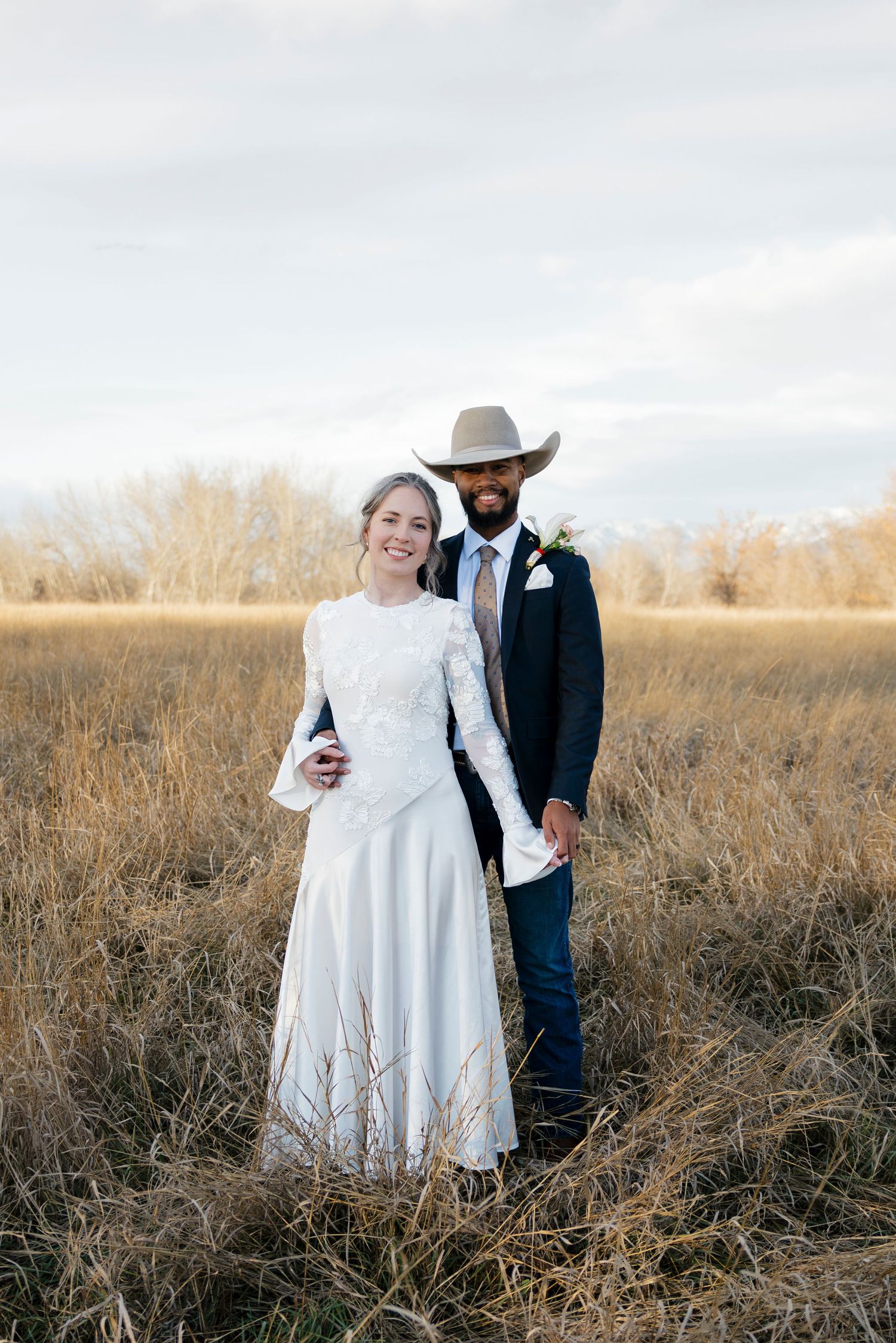Wedding couple in western attire posing in autumn field with mountains behind.