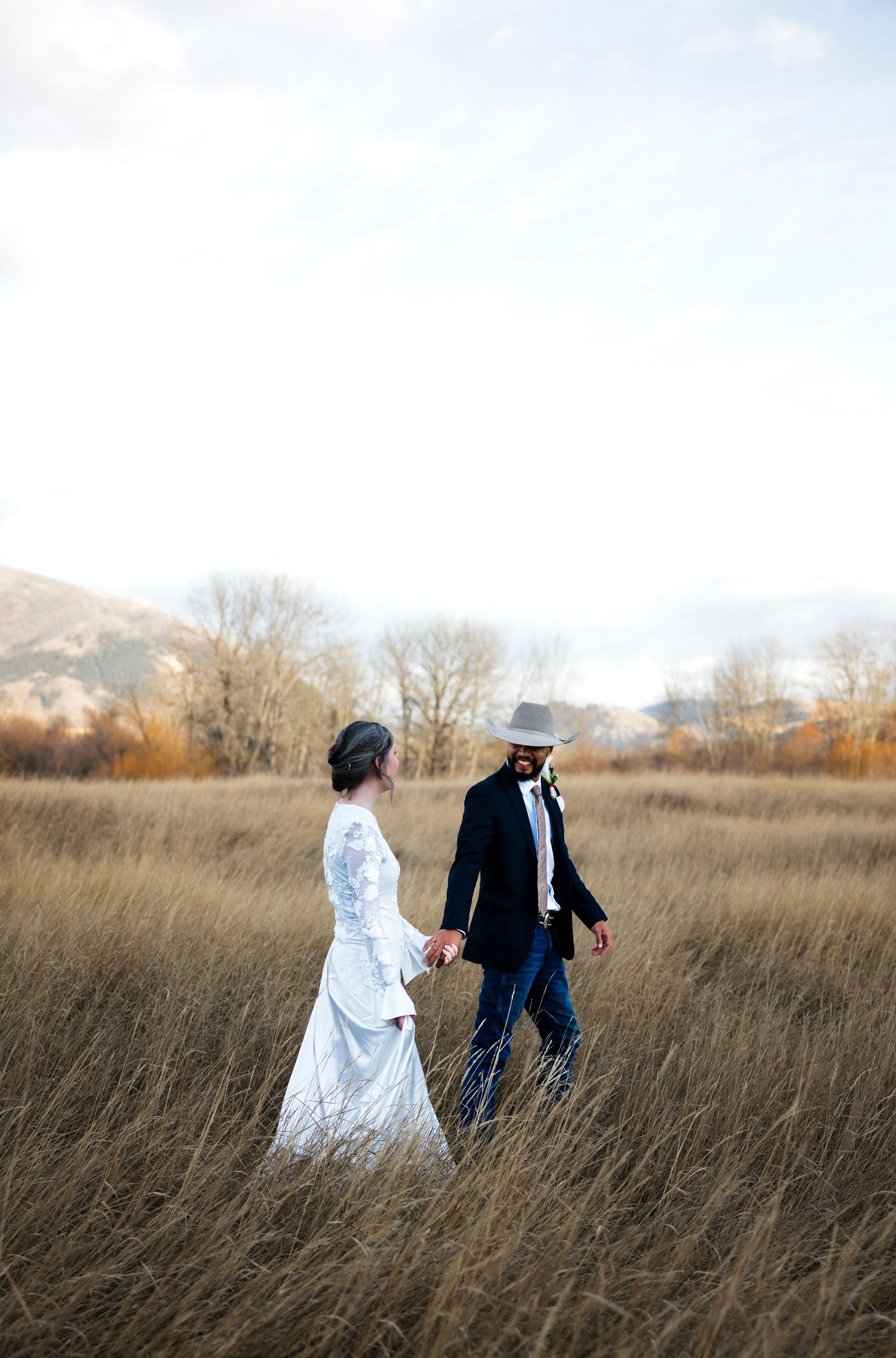 Couple walks through tall autumn grass in formal wedding attire during sunset.