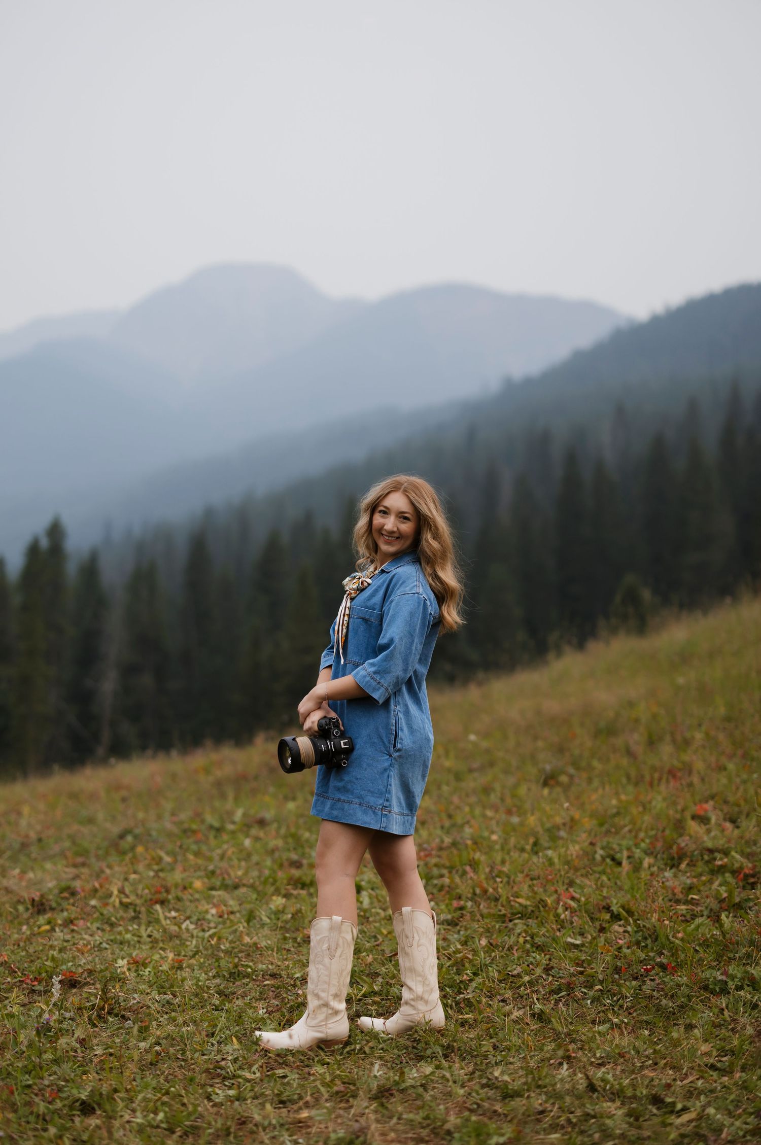 Young woman in denim dress and white cowboy boots holding a camera, smiling in a mountain meadow.